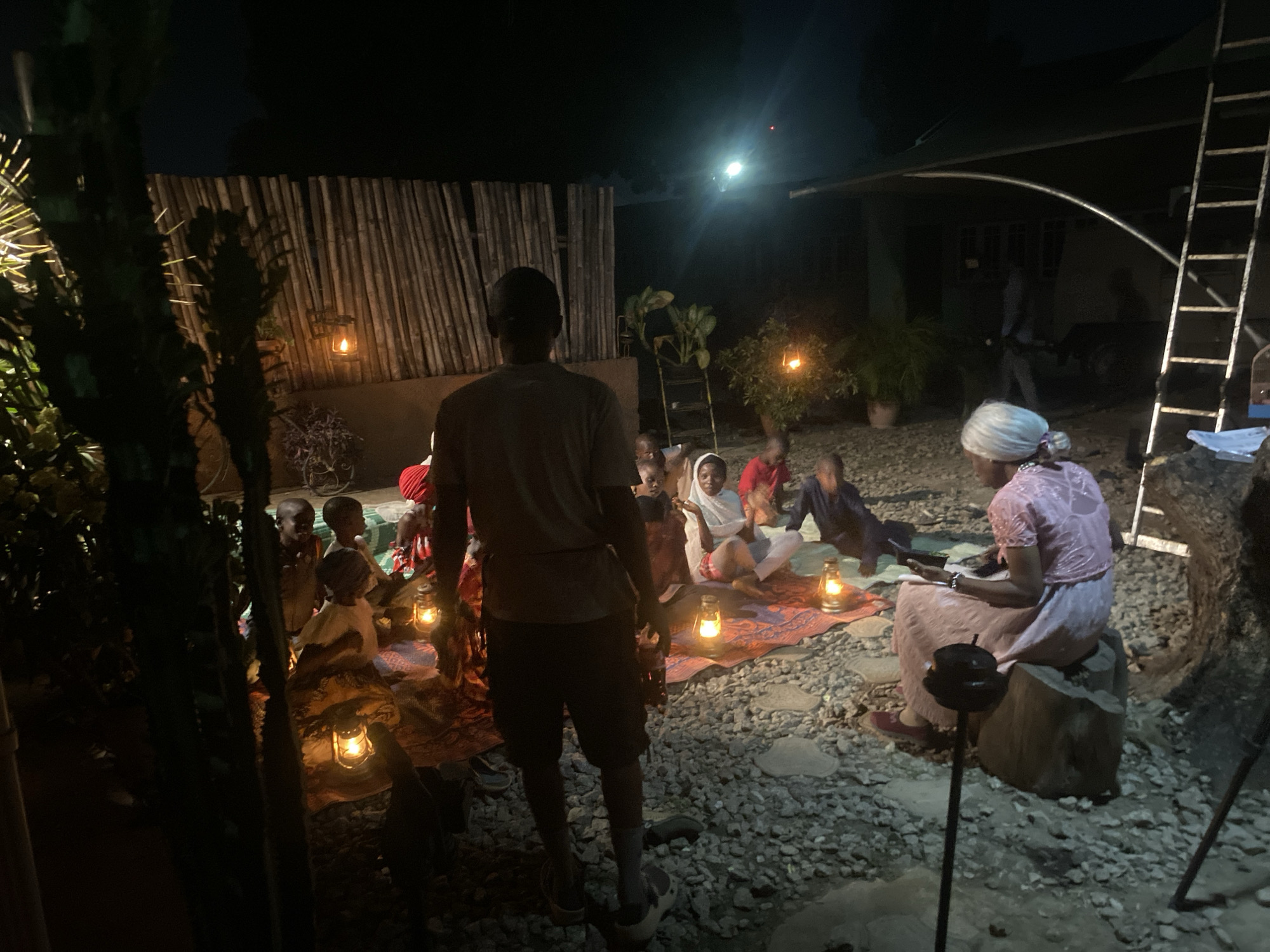 The image shows a group of children gathered around a seated older woman, possibly in Barden Yana, Nigeria, though this is not confirmed in the image itself. They are on a rough, stony ground, likely an outdoor courtyard. The setting is night, illuminated by several kerosene lamps placed near the children and one hanging on a bamboo-like fence. A man stands in the foreground facing the group. The woman is seated on a wooden stump, wearing a light-colored dress and a head covering. She appears to be reading from a book or tablet. A ladder leans against a building in the background. A person is walking near that building. The atmosphere is informal and intimate.