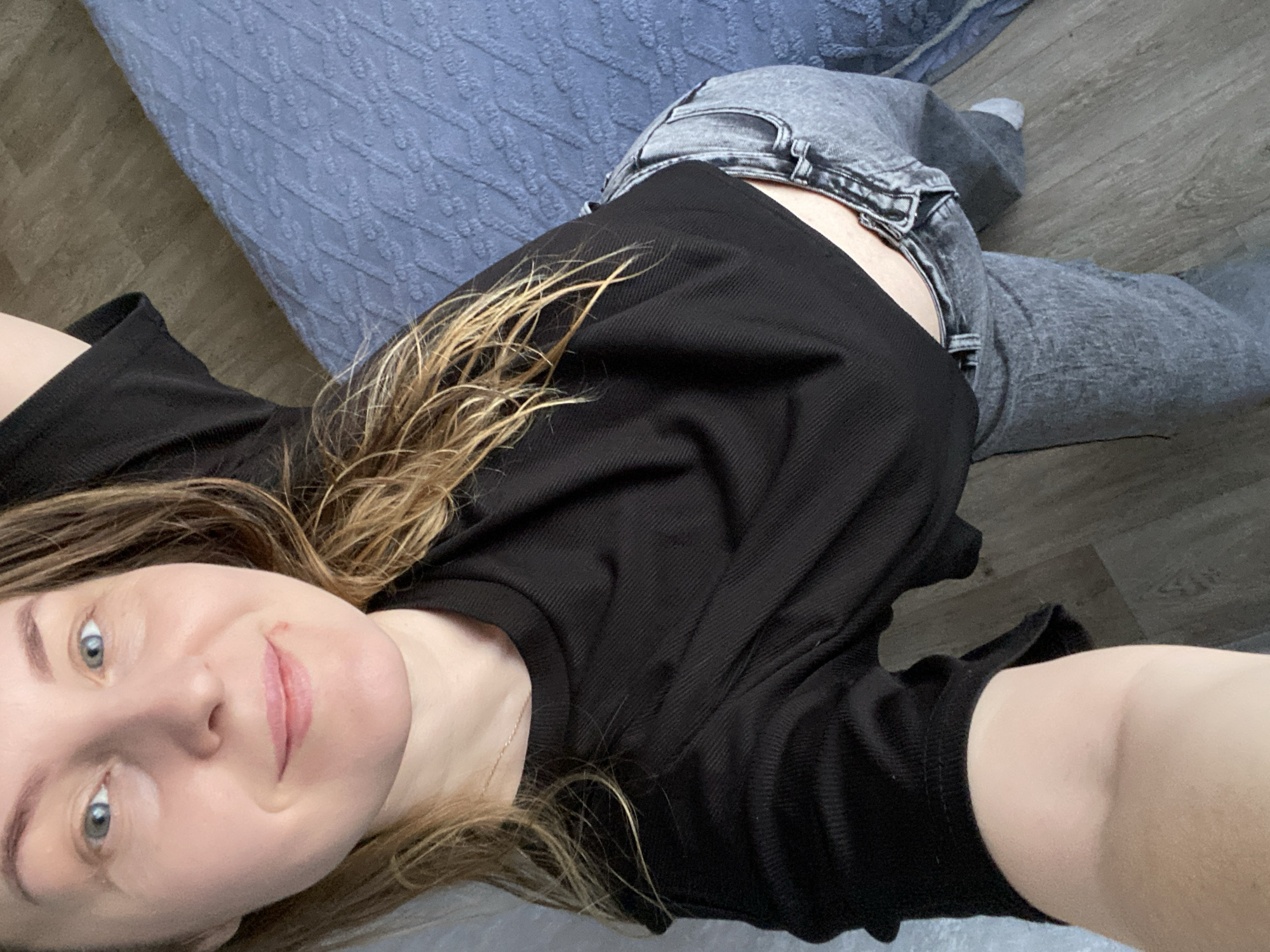 A woman lies on a surface, possibly a bed or a floor, wearing a black long-sleeved shirt and gray jeans. The surface appears to have a gray or blue patterned covering on one side, and a wood-look surface on the other. The woman is looking at the camera with a slight smile. Her hair is blonde and brown. The image does not indicate the location is Noviy, Russia.