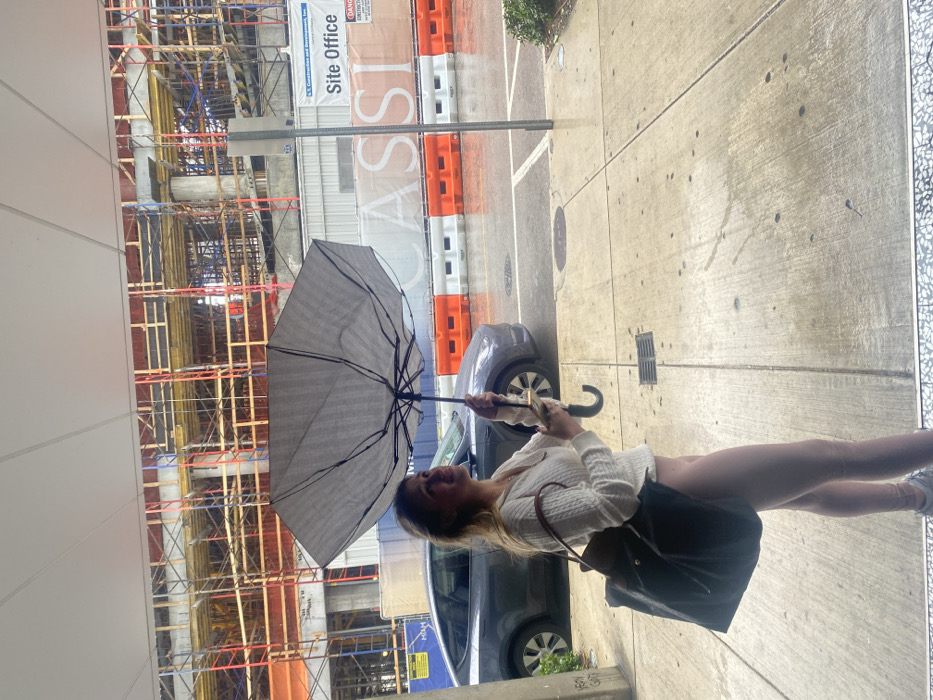 A woman holds an umbrella on a wet sidewalk. Behind her, a dark grey car sits next to a construction site with scaffolding. Orange and white barriers line the back of the sidewalk, and a sign reading "Site Office" is visible. The woman is wearing a white sweater, a dark skirt, and sneakers. She is holding the umbrella open with both hands. The image appears to be taken from a slightly elevated position.