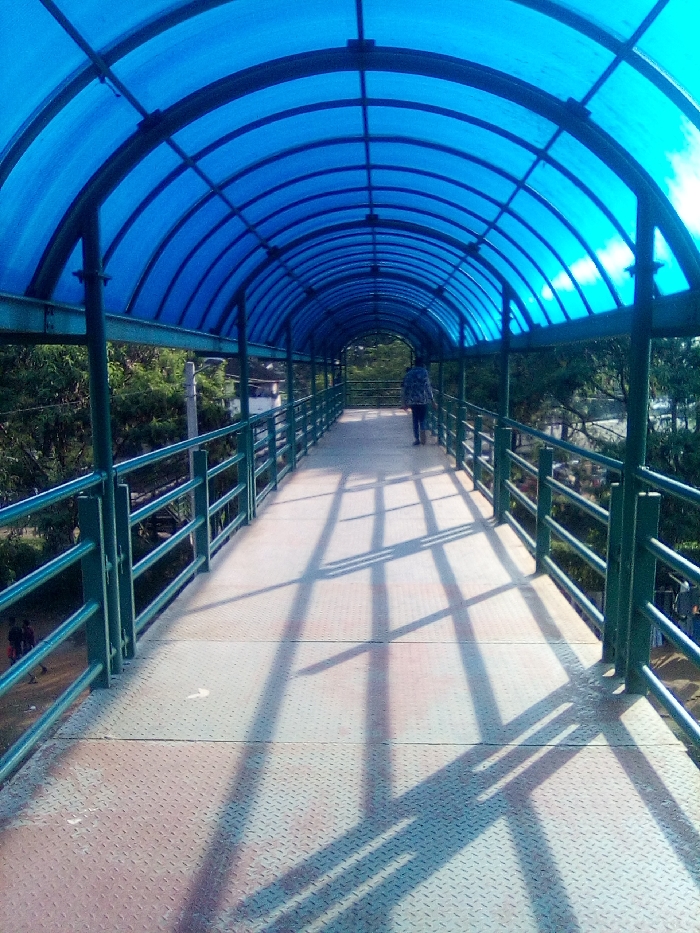 The scene depicts a pedestrian overpass in Bondo, Kenya. The overpass features a long, arched, translucent blue polycarbonate roof supported by dark green metal framework, extending into the distance. Below the roof, the walkway consists of gray, diamond-plate metal flooring. Dark green metal railings with multiple horizontal bars line both sides of the walkway. Strong sunlight penetrates the blue roof, casting distinct, angular shadows of the structural framework and railings onto the walkway surface. One individual, dressed in a patterned light-colored top and dark trousers, is observed walking away from the camera, toward the far end of the covered bridge. A white directional arrow is present on the walkway in the foreground, pointing forward. Below the bridge, on the left side, dense green foliage is visible, and faint figures of two or three individuals are partially discernible at ground level. The surrounding environment consists of abundant green trees and clear skies.