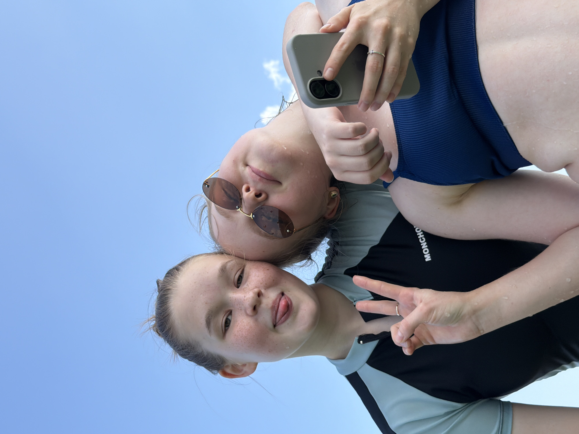 Two young females are pictured in a selfie outdoors against a clear blue sky. The female on the left, with freckles and her tongue sticking out, wears a black and grey rashguard. Her right hand is raised, making a peace sign. The female on the right, wearing sunglasses and a dark blue ribbed bikini top, leans on the other's shoulder. She holds a grey smartphone in her right hand. Both subjects appear to have water droplets on their skin. The logo "MONCHO" is visible on the rashguard of the female on the left. The background suggests an open, possibly coastal or waterfront, environment, characteristic of outdoor leisure activities. The image does not contain information that specifically places it in the city of Krasny Bor, Russia.