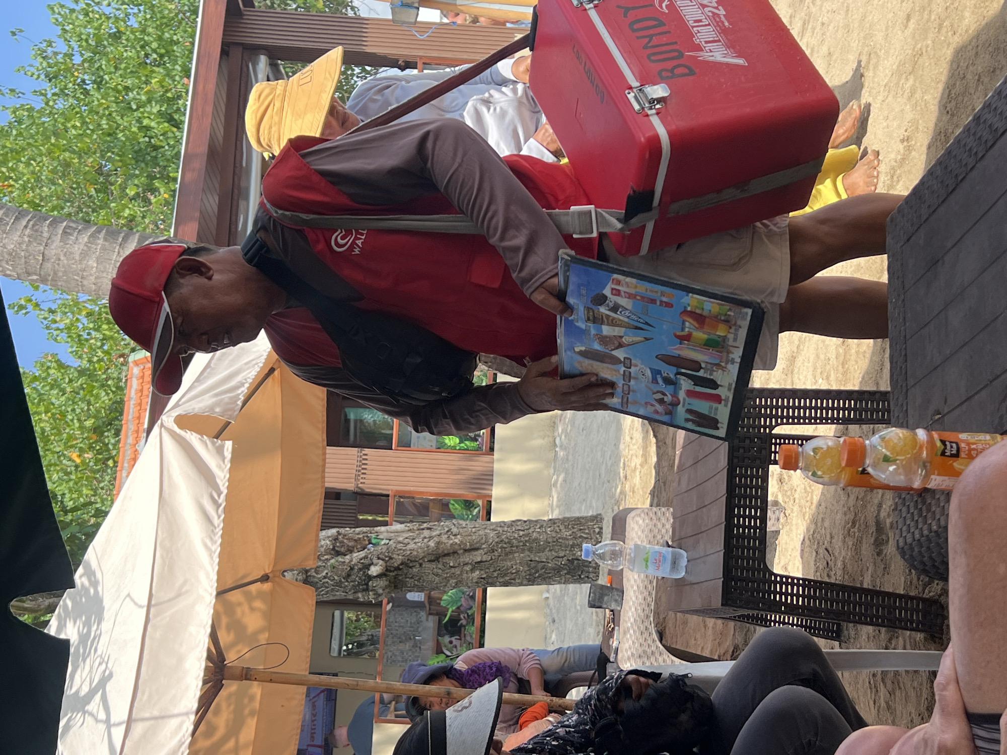In Kuta, Indonesia, on a sandy beach area, a man wearing a red cap and vest stands under an orange and white umbrella. He is holding a display board featuring various ice cream popsicles. To his right, a large red cooler with "BONDY" and "Marina Cooler 24" written on it is slung over his shoulder. Another person wearing a yellow hat is visible behind him. In the foreground, a brown plastic table holds two bottles of orange-colored liquid. The overall scene suggests a vendor selling refreshments on a beach.