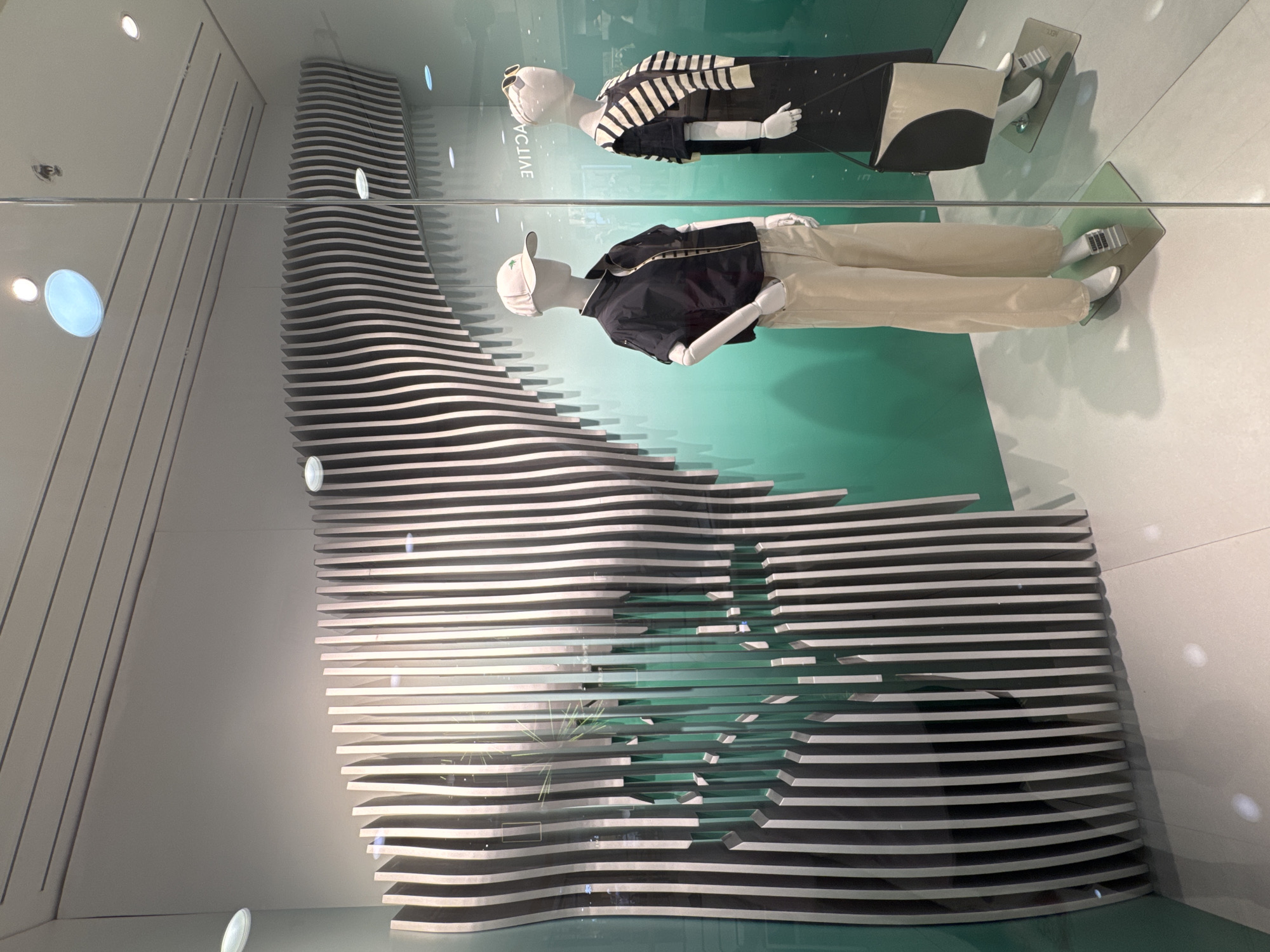 The image depicts a fashion store window display, likely a clothing store, with two mannequins. The display features a prominent, wavy, vertical slat design in black and white, creating a visually dynamic background. The mannequins are dressed in casual attire. The mannequin on the left wears a dark short-sleeved jacket, a white undershirt, light-colored wide-leg pants, and a white baseball cap with a green logo. The mannequin on the right is dressed in a black dress with a white and black striped knit sweater draped over its shoulders and is carrying a black and beige handbag. The background behind the mannequins transitions from a light grey at the top to a gradient of green towards the bottom. The floor of the display is a light gray tile. The scene is viewed from outside the window, with reflections visible on the glass. The store name "ACTIVE" is visible in reverse on a sign in the background. The setting is a commercial display, and there are no people actively interacting within the scene. The image captures a fashion display and does not contain information to determine the specific location in Linghe, China.