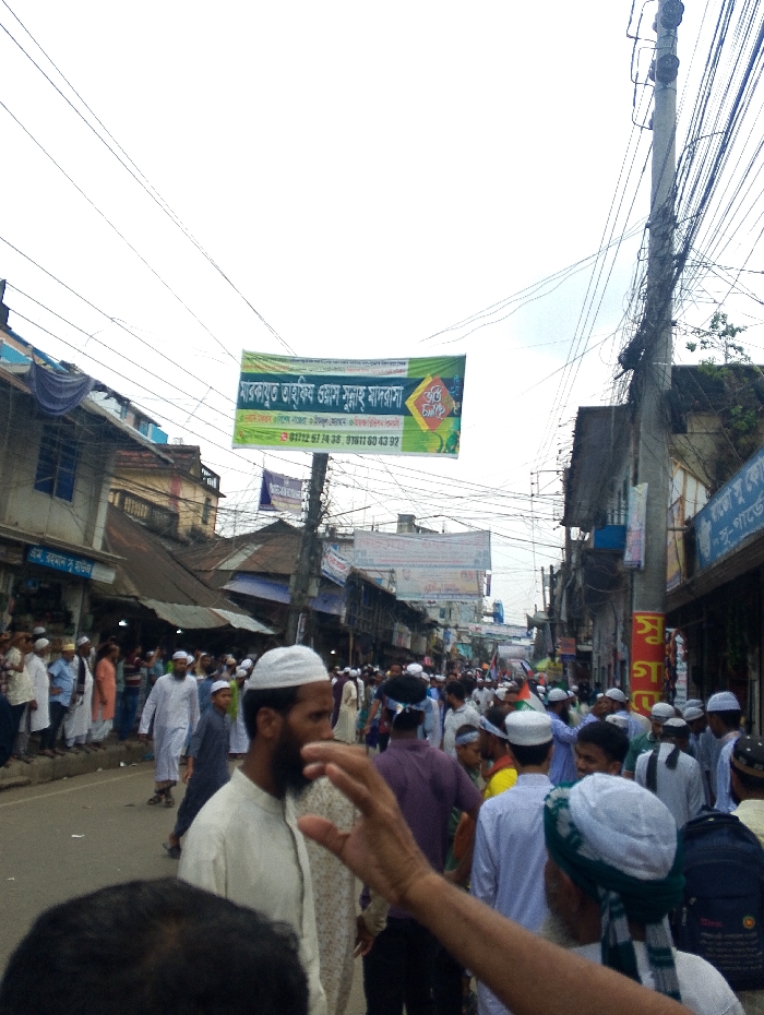 The image depicts a bustling street scene in Bhola, Bangladesh. The location is identified by its coordinates (latitude: 22.686, longitude: 90.644). The street is crowded with people, predominantly men, many of whom are wearing traditional white garments. There is a large banner hanging above the street, written in Bengali. Several shops are lined along the sides of the street, with a variety of goods displayed. An electric pole with numerous cables extends overhead, adding to the chaotic yet lively atmosphere of the scene. The focus is on the crowd of people, their movement, and the overall ambiance of the street.