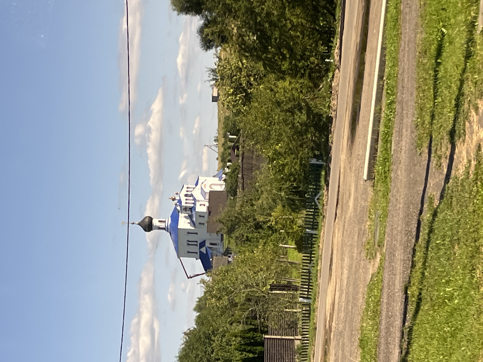 The image depicts a view of a white church with a blue dome and a black dome, located on a hill. The church is surrounded by trees and some buildings. In the foreground, there is a dirt road with some grass on the sides and a wooden fence. The sky is blue with scattered white clouds. There are no people or animals visible in the image. The overall scene suggests a rural or suburban setting. Based on the architectural style of the church and its surrounding environment, the image likely shows the Church of the Exaltation of the Holy Cross in Myadzyel, Belarus.