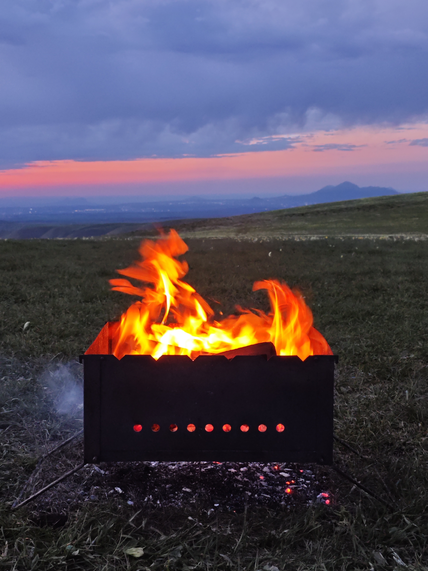 A charcoal grill sits on a grassy hill in an outdoor setting during twilight. The grill is filled with burning wood or charcoal, producing bright orange and yellow flames. Smoke rises from the side of the grill. In the background, a landscape stretches out with a distant city skyline visible under a pink and purple-tinged sky with clouds. The environment suggests a rural or elevated location overlooking the city.