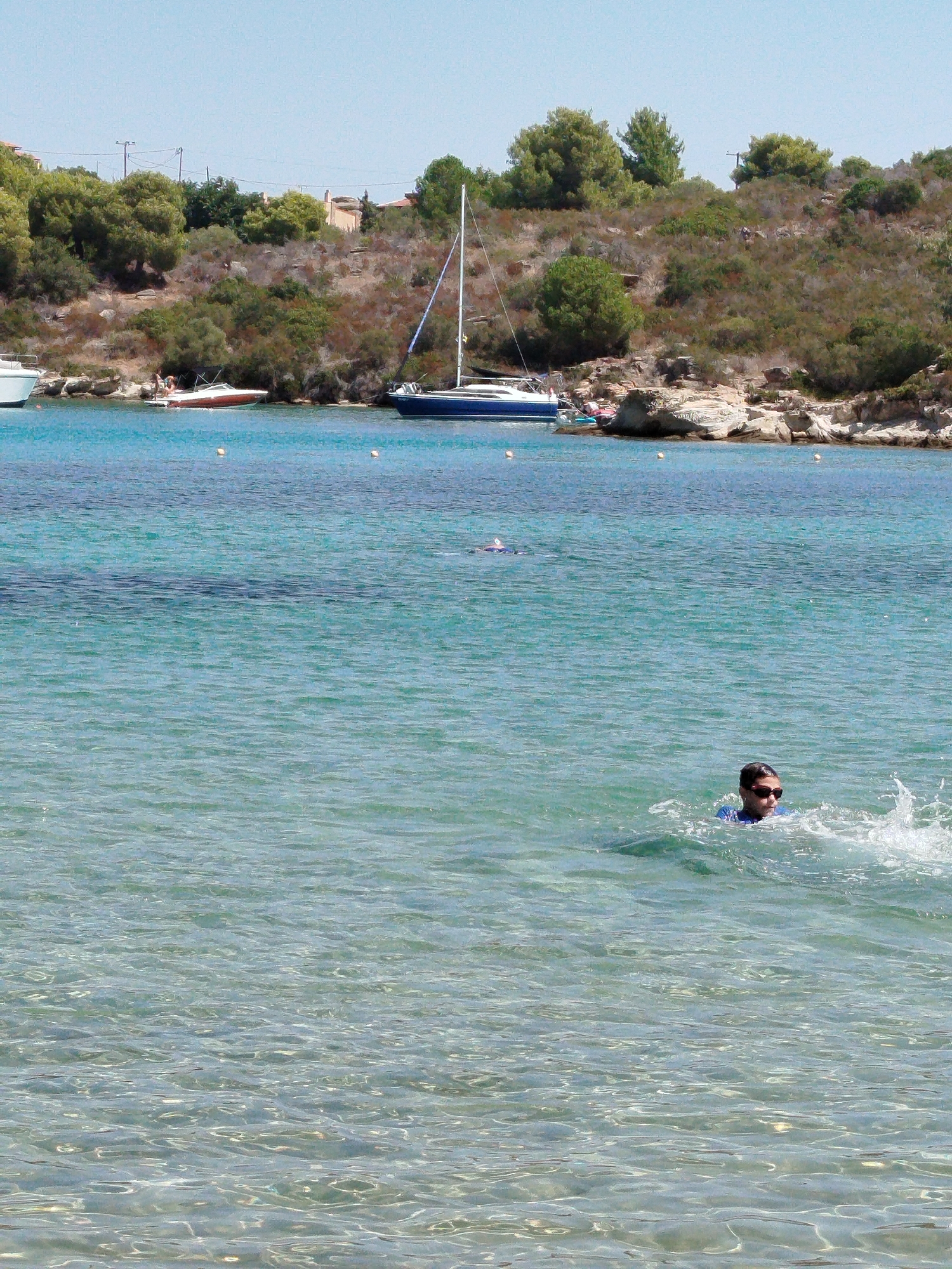 The image shows a scenic view of the sea in Sithonia Municipal Unit, Greece. In the foreground, a person is swimming in the clear turquoise water, wearing sunglasses and a blue shirt. The water is calm with gentle ripples, and sunlight reflects off the surface, creating bright spots. In the middle ground, there are several boats moored near the shore. One is a white and red speedboat with people on board, and a larger blue sailboat is anchored further out. The background features a rocky, tree-covered hillside under a clear blue sky. The overall impression is a peaceful day at the beach.