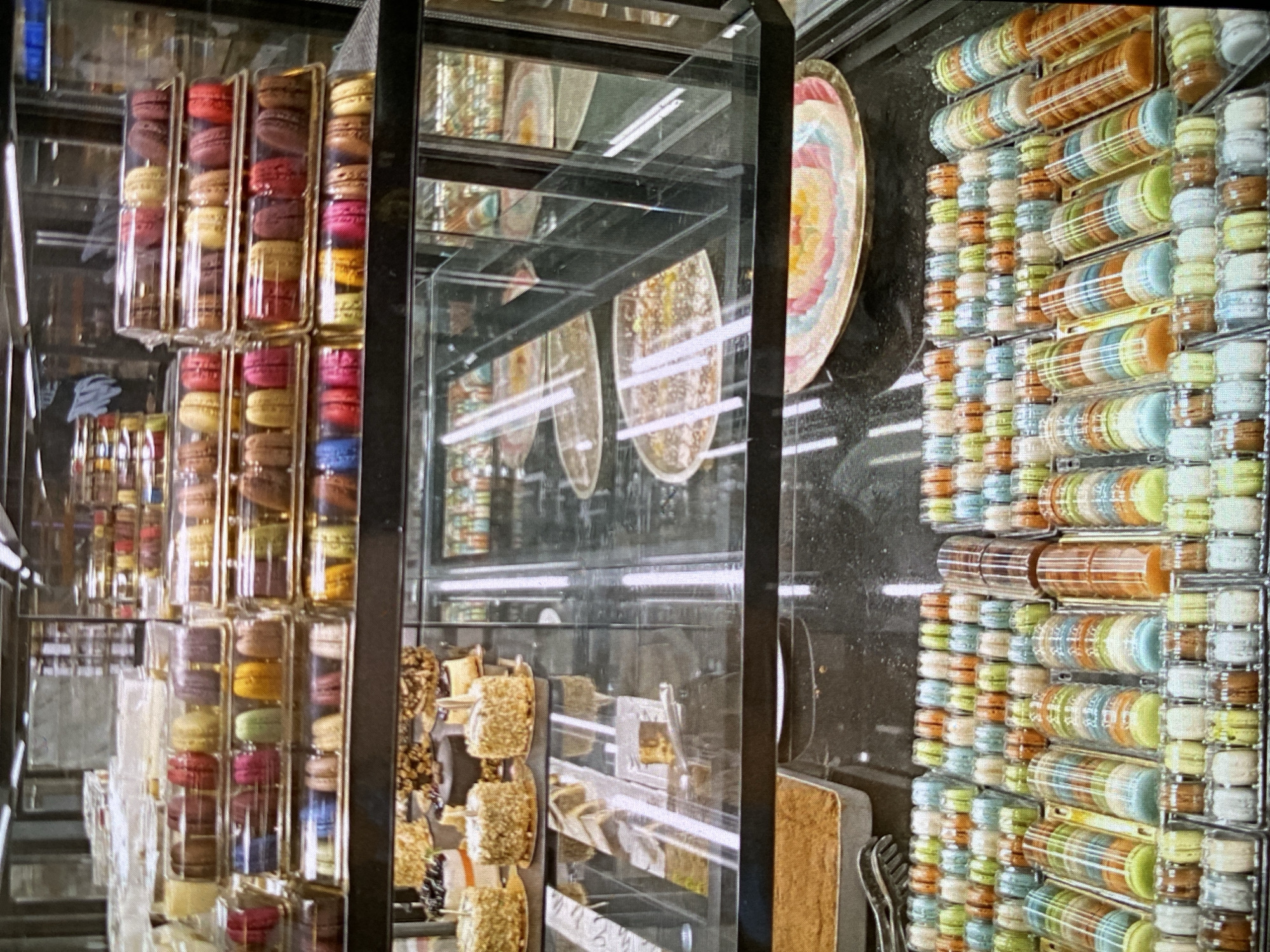 This image presents a vibrant, colorful display of macarons within what appears to be a retail setting, likely a dessert shop in Bankya, Bulgaria. The main subjects are numerous macarons, meticulously arranged in clear plastic packaging inside a glass display case.
On the left side of the frame, macarons are stacked vertically in rectangular compartments within larger clear boxes, showcasing a spectrum of colors including red, pink, yellow, green, brown, purple, and blue. On the right, and extending vertically, are long, clear cylindrical tubes filled with macarons, stacked horizontally. These tubes contain macarons in softer pastel shades of blue, green, orange, light brown, and white. Below these tubes, additional macarons are arranged in small, individual round clear containers, forming a dense, colorful grid.
The primary activity depicted is the merchandising and display of these confections for sale. The image is taken through glass, which creates prominent reflections that add visual complexity. Notable reflections include a large, round decorative plate featuring concentric pastel bands of pink, orange, yellow, green, and blue, as well as reflections of other patterned surfaces and bright artificial light sources appearing as distinct horizontal white streaks. Through the reflections and glass, other sweet items like what appear to be candied apples or similar confections coated in nuts on sticks, and silver tongs, are partially visible.
No people are present in the image. The time of day is not explicitly discernible, but the scene is brightly illuminated by artificial lighting, indicating an indoor commercial environment during operational hours. There is no legible text visible on any of the products or packaging. The overall scene conveys a sense of rich variety and appealing presentation of desserts.