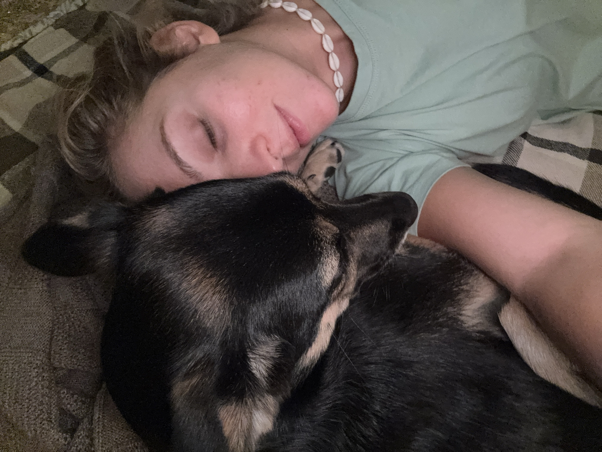 A woman is sleeping with a dog. The woman is lying on her side, with her eyes closed. She is wearing a light green t-shirt and a shell necklace. The dog is lying close to her, with its head resting near her face. The dog is primarily black with light brown patches. They are both lying on a bed with a plaid patterned bed cover. The location cannot be determined as being in Minsk, Belarus from this image.