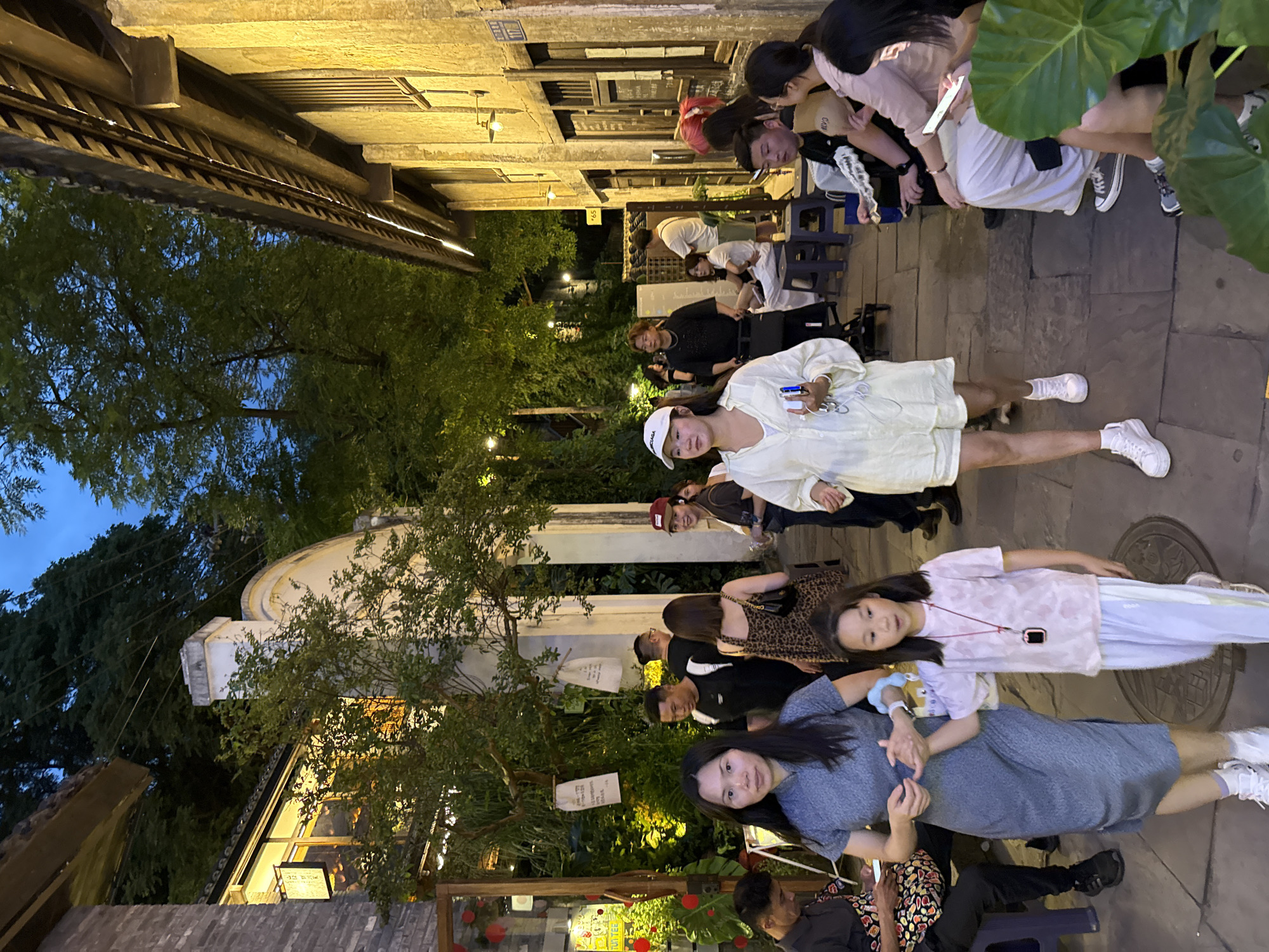 A nighttime scene in Nan'an District, China, shows a narrow cobblestone street illuminated by warm lighting from buildings and strung lights. A diverse group of people are gathered, primarily seated on small stools, suggesting an outdoor dining or social setting. Individuals appear to be engaged in conversation or waiting, some glancing at the camera. Plant life is visible throughout, adding to the ambiance, and the architecture is a mix of traditional and modern styles.