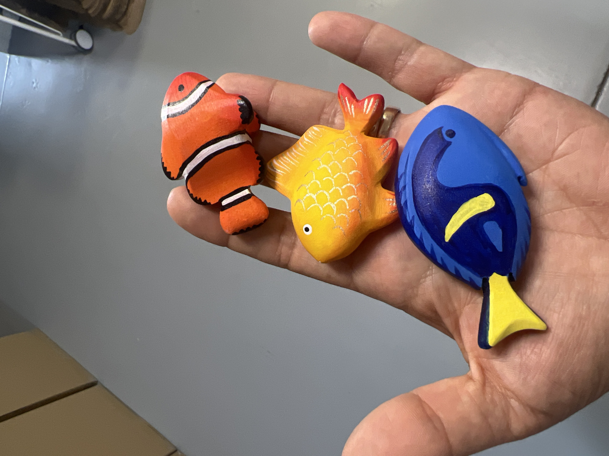 A person's hand holds three painted wooden fish. The fish are small, decorative objects, possibly magnets or charms. From left to right, the first fish is orange with white and black stripes, resembling a clownfish. The second fish is yellow with a textured pattern of scales and red accents on its fins. The third fish is blue with a yellow stripe and black details, resembling Dory from "Finding Nemo". The hand is lightly cupped, showing the palm and fingers. A ring is visible on the ring finger. The background is out of focus and appears to be an indoor setting with a light gray wall and a portion of a light brown surface. The lighting suggests an indoor environment, possibly during the daytime. No text is visible in the image.