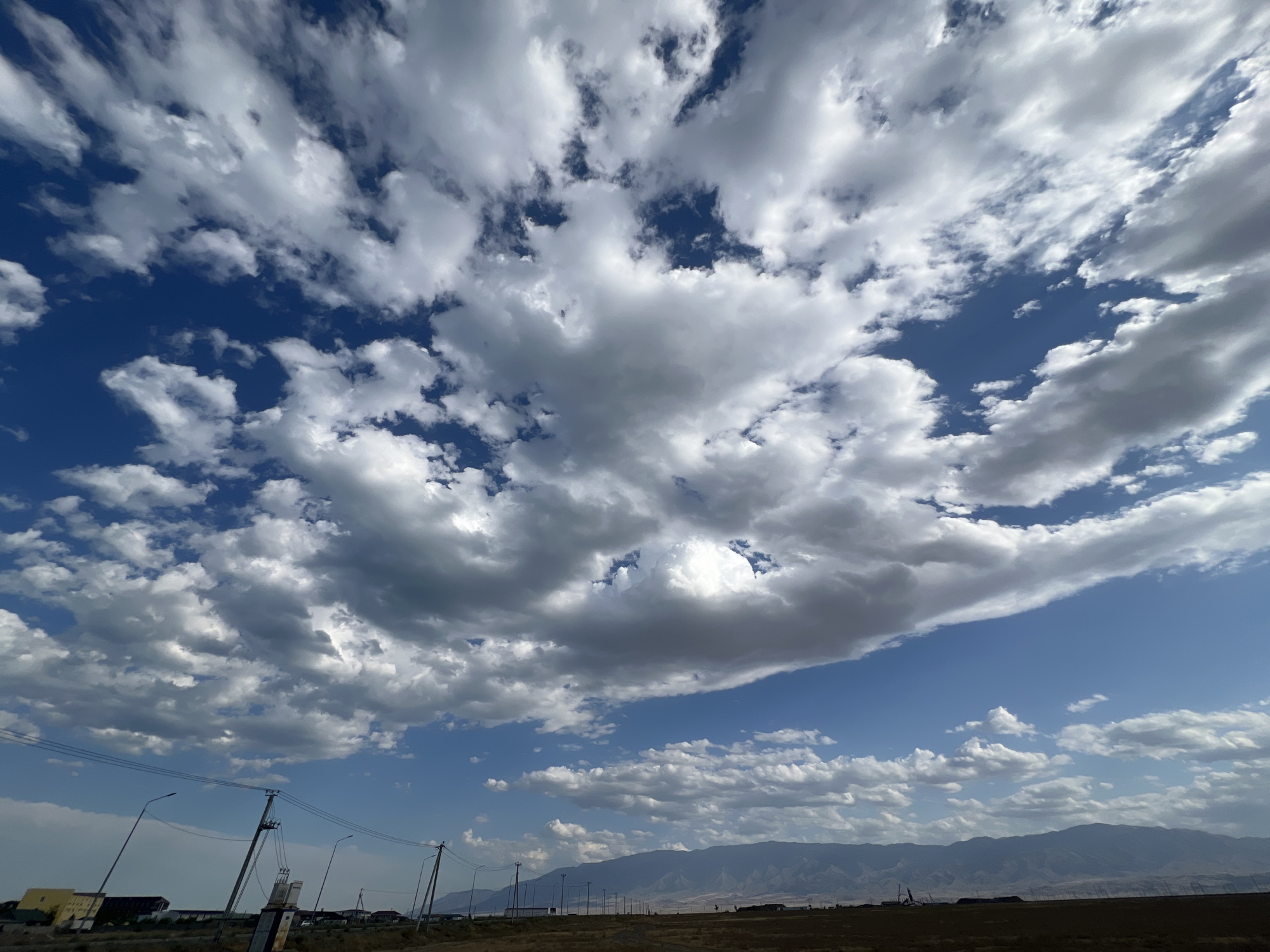 The image captures a wide, low-angle view of a dramatic sky filled with cumulus clouds, set against a clear blue sky. The clouds are varied in density and shape, with some appearing bright white and puffy, while others are darker and more elongated, suggesting varying altitudes and moisture content. The sky occupies the majority of the frame, emphasizing its grandeur.
In the lower portion of the image, the horizon is visible, showing a flat, arid landscape with sparse vegetation. A power line with several poles and wires stretches across the middle ground, indicating a rural or semi-urban setting. In the far distance, a range of mountains is silhouetted against the sky, adding depth to the scene. On the left side, a few buildings are discernible, including what appears to be a yellow structure and some smaller residential or industrial buildings. The overall impression is one of open space and natural beauty, with signs of human infrastructure present but not dominant. The location is identified as Ақши, Kazakhstan.