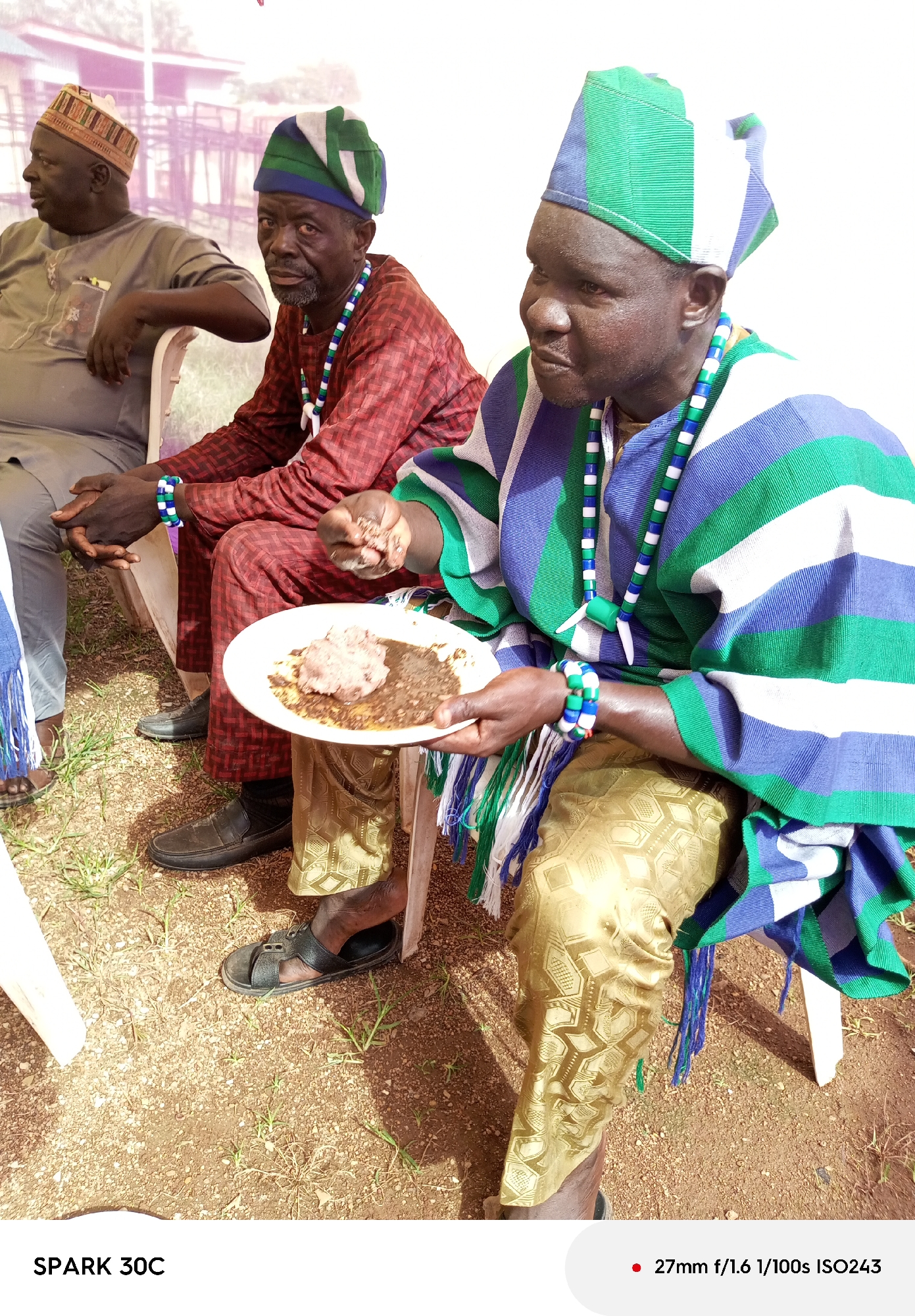 Three men sit on white plastic chairs outdoors on gravel-covered ground, with some grass visible. All wear traditional clothing and headwear. The man closest to the camera wears a green, white, and blue striped tunic, gold pants with patterned designs, and a matching hat, and has a beaded necklace and bracelet. He holds a white plate with a brown, dough-like food item and dark sauce. The man in the center wears a red and black checkered outfit with a green, white, and blue hat, with a beaded necklace and bracelet. He sits with his hands clasped in his lap. The man on the left wears a gray outfit with a brown and patterned hat and is seated with his hands resting on his knees. The location is identified as Maigizo, Nigeria.