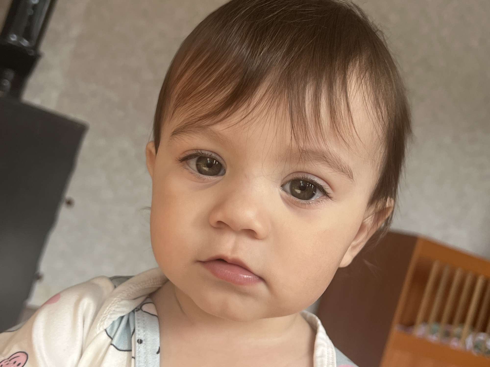 A close-up shot features a young child with brown hair and brown eyes. The child's skin tone is light. The child is wearing a patterned white and blue top. A dark object is partially visible in the background, to the left of the child, and a wooden object is visible to the right. The background is a plain, neutral color.
The location information is not discernible in the image.
