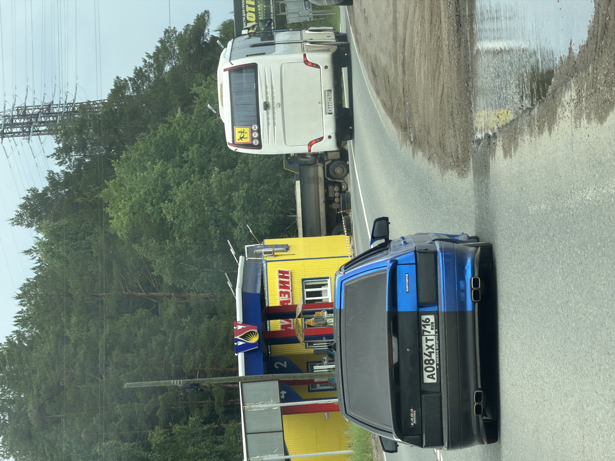 This image captures a roadside scene in what is identified as Noviy, Russia, under an overcast sky.
In the foreground, a two-tone Lada Samara automobile is driving away from the viewer. The car's left side is black, and its right side is a vibrant blue, with a distinct dividing line running vertically down the center of its rear. It bears Russian license plate A084XT 716, indicating the Ulyanovsk Oblast region. The car features "Lada Sport" and "Samara" badging, along with a rear spoiler and quad exhaust tips (two on each side).
Further down the asphalt road, a white passenger coach bus with Russian license plate P777YB 52 (Nizhny Novgorod Oblast region) is also moving away. The bus displays yellow square decals on its rear window: one depicts two children, and others show speed limits of 60, 70, and 80 km/h.
To the left of the road, behind the Lada, stands a distinctive yellow building with blue roof accents and red structural columns. A prominent V-shaped logo with a yellow and red emblem is on the facade. The building has the Russian word "МАГАЗИН" (MAGAZIN, meaning "Shop") in red lettering on its side, along with the numbers "2" and "4" on two of its columns. A small, open-air umbrella structure is situated in front of the shop's entrance. A brown wooden utility pole is visible to the left of the shop.
Behind the bus and to the right of the shop, a dark-colored heavy-duty truck with a flatbed or low-side bed is partially visible, appearing to be stationary off the main road.
The background is dominated by dense green foliage, primarily coniferous trees (pines) and some deciduous trees, indicating a forested or semi-rural environment. High-voltage power lines and a large metal lattice pylon are visible against the pale, overcast sky in the upper right quadrant of the image. The road has a rough, gravelly shoulder, and a large puddle reflecting the sky and parts of the environment is present on the right shoulder in the mid-ground.