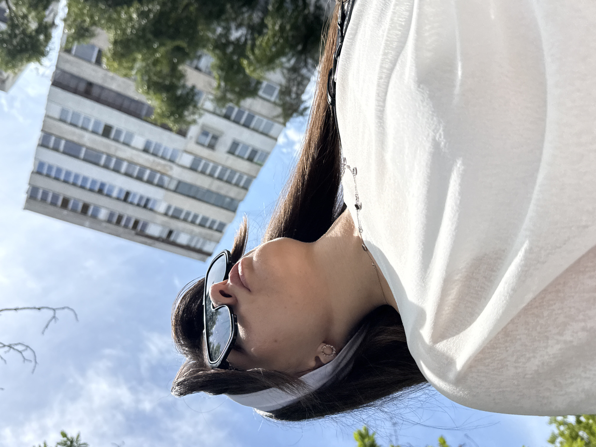 A low-angle, close-up shot shows a person wearing sunglasses and a white headscarf, looking upwards. The person has long, dark hair and is wearing a white, loosely draped top. A silver necklace with small dark pendants is visible. In the background, a large, multi-story, light-colored building with many windows is partially obscured by green trees. The sky is blue with scattered clouds.