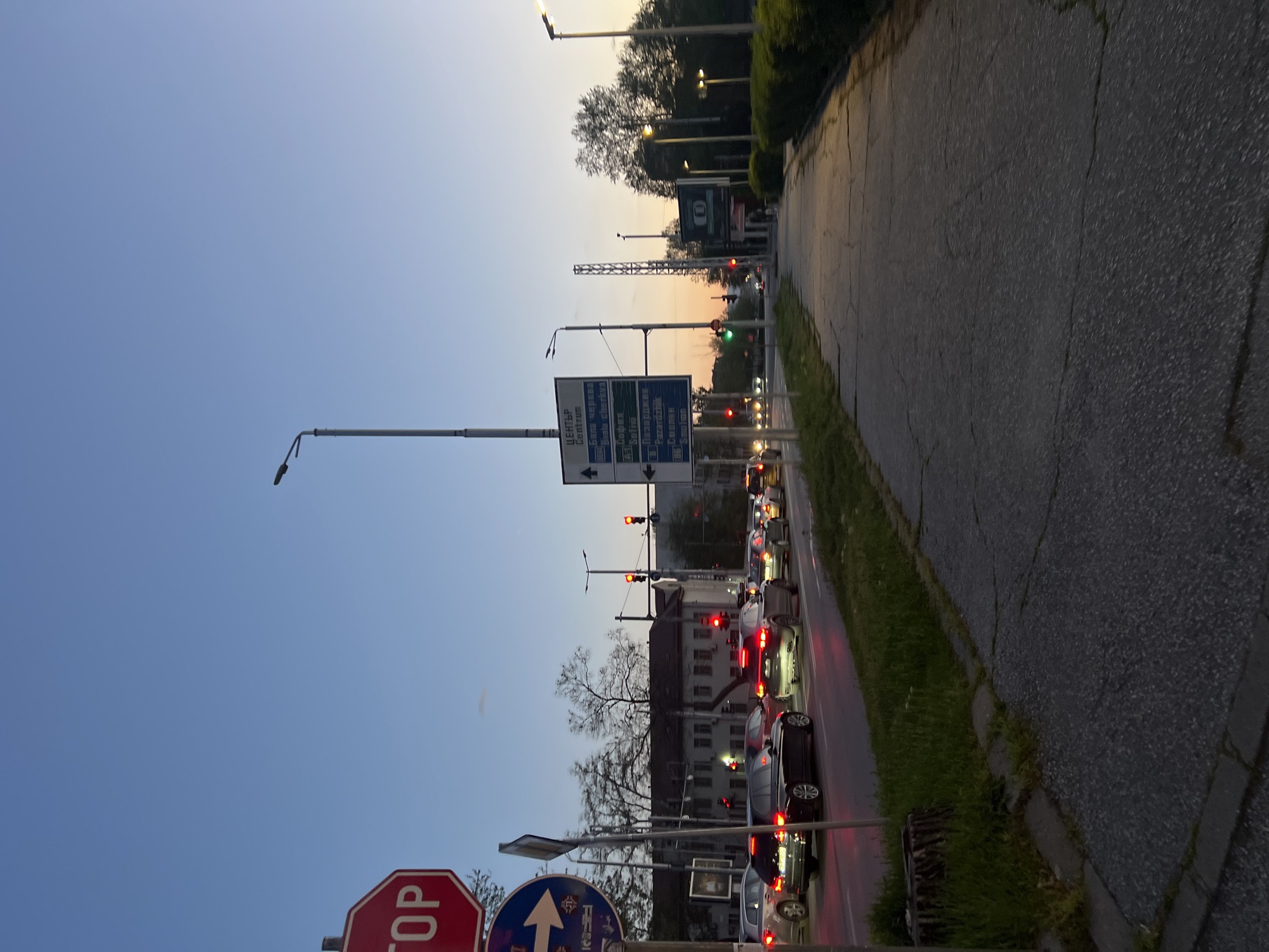 This media file depicts an urban street scene in Plovdiv, Bulgaria, captured during dusk or early evening. The sky, transitioning from a soft blue at the top to a warm orange-yellow near the horizon, indicates the setting sun or twilight hours.
The main subjects include a busy road filled with queued vehicles and various street signage. Numerous cars are visible on the street, with their red taillights illuminated, suggesting they are stopped at a traffic light or experiencing heavy congestion. Several traffic lights are also present, some showing red and others green.
On the left side of the frame, prominent road signs guide traffic. A large multi-panel directional sign displays destinations in both Cyrillic and English, reading: "ЦЕНТЪР Centrum", "Пловдив Северна Промишлена Зона", "А-1 София", "Пазарджик", "Смолян", and "Самоков", accompanied by directional arrows. Below this, a red octagonal STOP sign is partially visible, along with a blue circular "one-way" sign featuring an upward-pointing arrow.
To the right of the road, a sidewalk or pedestrian path, characterized by cracked asphalt, runs parallel to the street. It is bordered by a strip of green grass and several bushes and trees. Streetlights are illuminated along the roadside. In the mid-ground, behind the traffic, several multi-story buildings, typical of an urban setting, are visible. No people are explicitly seen, only their vehicles in transit or stopped. The scene captures a moment of typical urban activity as the day turns into night.