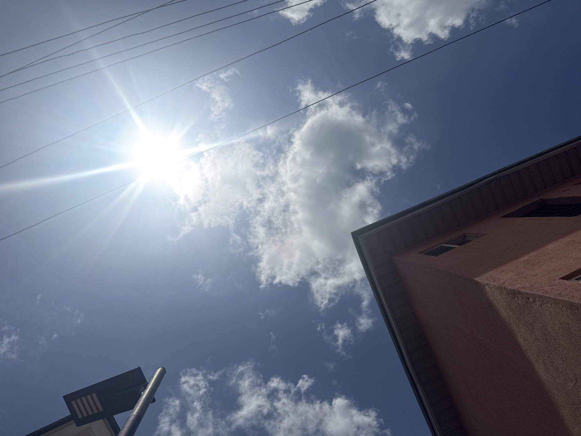 The image, taken from a low angle in Ihiagwa, Nigeria, shows a bright, sunny sky with scattered cumulus clouds. A set of power lines runs diagonally across the upper part of the frame, intersecting the sun. A section of a reddish-brown, multi-story building is visible in the lower-right corner, with a few visible windows. In the lower-left corner is a portion of a streetlight and its pole.