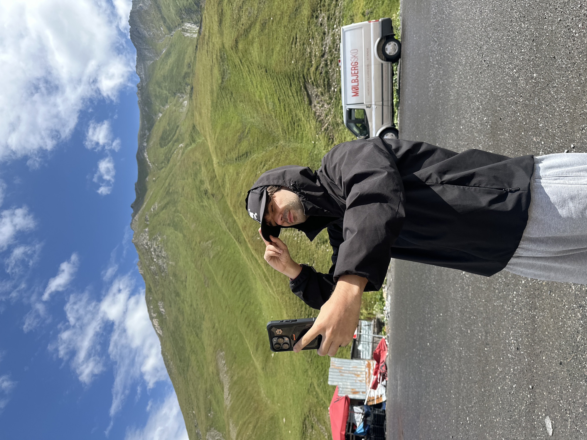 A man in a black hooded jacket and a baseball cap takes a selfie with his phone. He is standing on an asphalt surface with a green, mountainous landscape in the background under a blue sky with white clouds. To his left, there are some temporary structures and a red umbrella, and further in the background, a white van with the text "MØLBIERGSKO" is parked. The location appears to be a rural mountain pass, potentially in the Almasiani region of Georgia, given the terrain and style of the van.