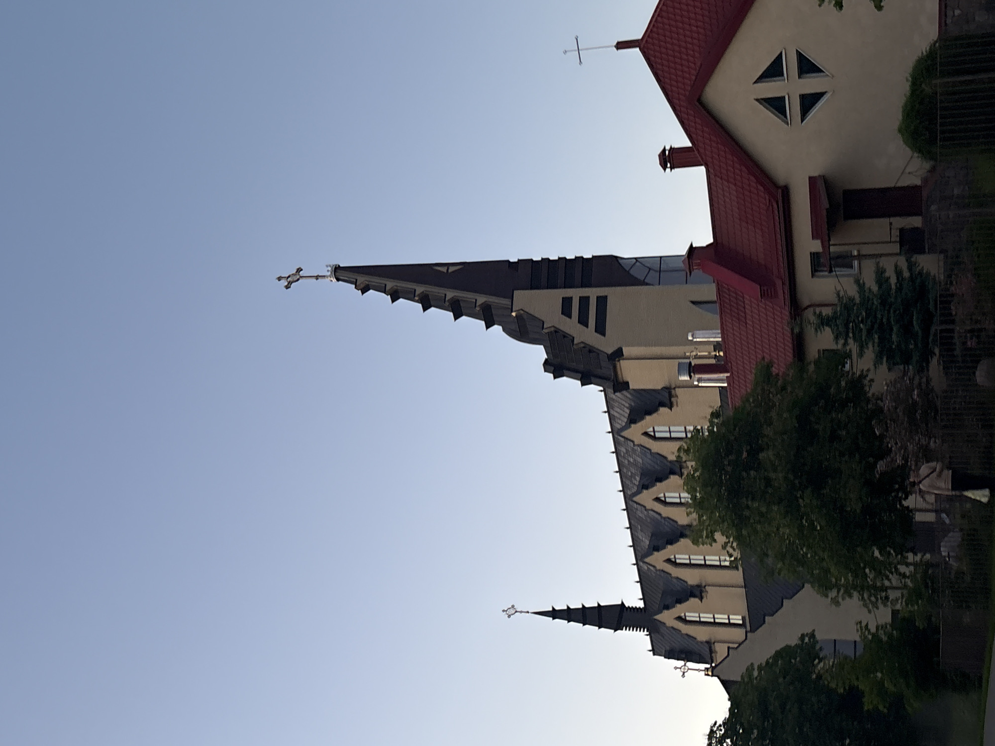 The image shows a view of a church in Minsk, Belarus. The church has a tall, pointed spire topped with a cross, as well as a building with a red roof and triangular windows attached to it. The sky is light blue. Part of the church is obscured by trees.