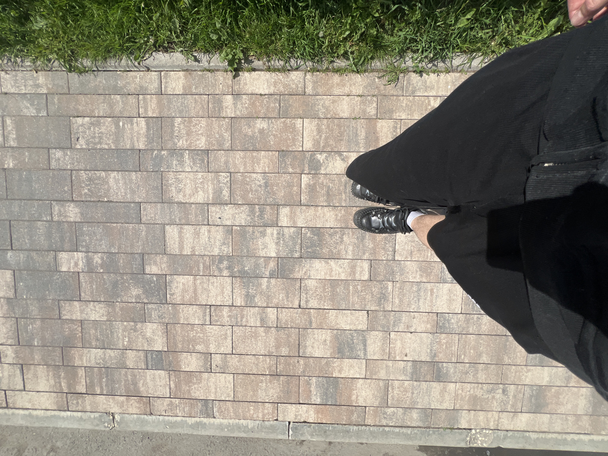 A top-down view of a person's legs and feet standing on a paved walkway in Saint Petersburg, Russia. The walkway is composed of rectangular interlocking pavers in various shades of brown and grey. To the right of the walkway is a strip of lush green grass. To the left of the walkway is a grey concrete curb and a patch of dirt. The person is wearing black pants, white socks, and black shoes. The sun is casting shadows on the pavers and the person's legs.