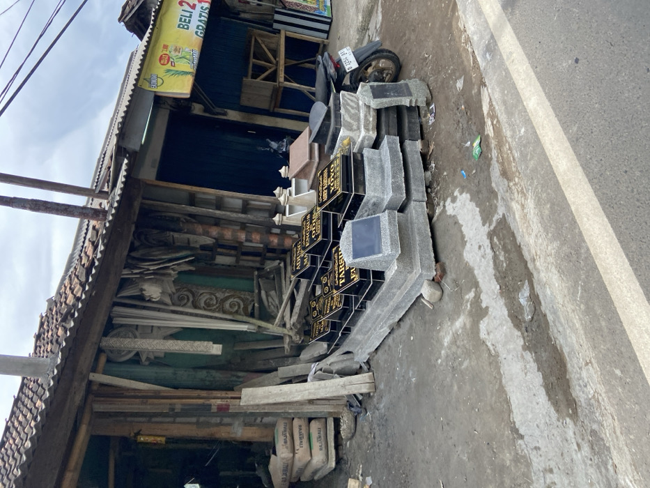 The image shows a street side storefront in Soreang, Indonesia (latitude: -7.033, longitude: 107.504). The storefront is selling gravestones, which are stacked against the building, and there are a few other objects, such as a motorcycle, and cement bags stacked nearby. The gravestones are black with gold lettering. The motorcycle has a license plate that says “D 2234 BB.” The building is old and dilapidated, with a corrugated metal roof. The sidewalk is paved in grey concrete and is bordered by a white painted line.  There is a small amount of debris on the sidewalk.