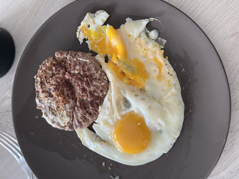A dark brown, round plate containing food is positioned on a light-colored, wood-grain surface in Chambœuf, France. The plate holds one cooked, browned meat patty, approximately circular, which exhibits a crack or separation across its center. Adjacent to the patty are two fried eggs. The egg on the left has an intact bright yellow yolk and firm white. The egg on the right has a broken yolk, with the liquid yellow yolk spreading across its white and onto the plate's surface. A silver-colored fork is partially visible in the upper left of the frame, and a dark, cylindrical object, possibly a cup or mug, is partially visible in the upper right. No individuals are present in the scene.