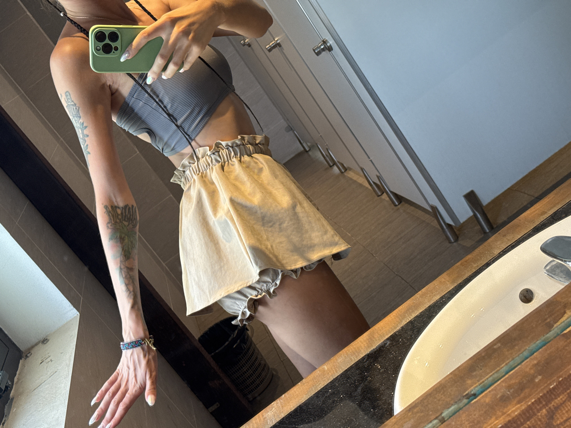 A person is seen taking a mirror selfie in what appears to be a public restroom, located in Hà Giang 1 Ward, Vietnam. The image captures a partial reflection of the individual, focusing on their torso, left arm, and upper legs.
The person is wearing a grey ribbed crop top and a light beige or tan high-waisted ruffled skirt, with ruffled undergarments or shorts visible beneath the skirt. They are holding a light green smartphone with a black camera module in their right hand, reflected as their left, positioned to capture the image. The phone has a black strap, possibly worn around the neck or shoulder. The person's left arm is prominently displayed, adorned with multiple tattoos: a detailed, tower-like structure with flowing vines or leaves on the upper arm, and a darker, less decipherable design on the lower forearm. A multicolored beaded bracelet with a small charm is visible on their left wrist. Their nails are long and manicured with a light, possibly iridescent, polish.
The background of the reflection reveals a row of several light-colored, closed stalls or cabinets, each with a silver handle or pull. In the immediate foreground, part of a white sink with a drain and a metallic faucet is visible, set into a dark countertop with a rustic-looking wooden frame around the mirror and sink area. A black wicker-like basket or trash can is faintly reflected on the floor beneath the skirt. The lighting suggests an indoor setting, likely daytime. No discernible text is visible on any objects or surfaces in the image. The activity is clearly a mirror selfie.
