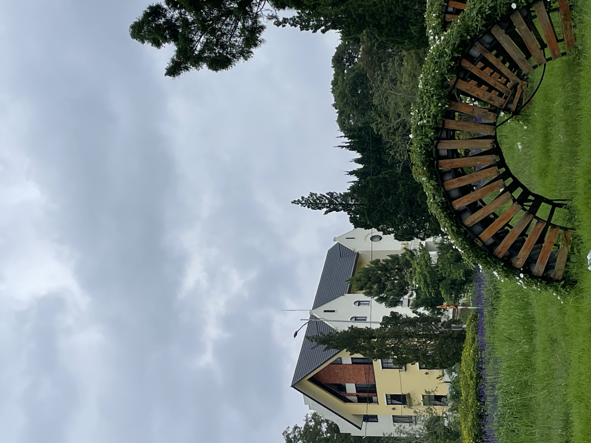 In the city of Da Lat, Vietnam, a picturesque scene unfolds under a cloudy sky. A charming, multi-story building with a distinctive yellow and white facade stands prominently, its architectural style suggesting a European influence with its pointed gables and dark, sloping roof. The building is partially obscured by lush green trees, including tall cypress-like evergreens.
In the foreground, a creatively designed garden archway constructed from weathered wooden planks and greenery creates a natural frame. This archway is adorned with small white flowers and lush green foliage, suggesting a well-maintained garden space. Beyond the archway, a field of vibrant green grass stretches out, interspersed with rows of purple flowers, likely lavender, adding a splash of color to the landscape. In the mid-ground, a row of yellow flowers adds further visual interest. The overall atmosphere is serene and tranquil, characteristic of Da Lat's many beautiful gardens and European-inspired architecture.