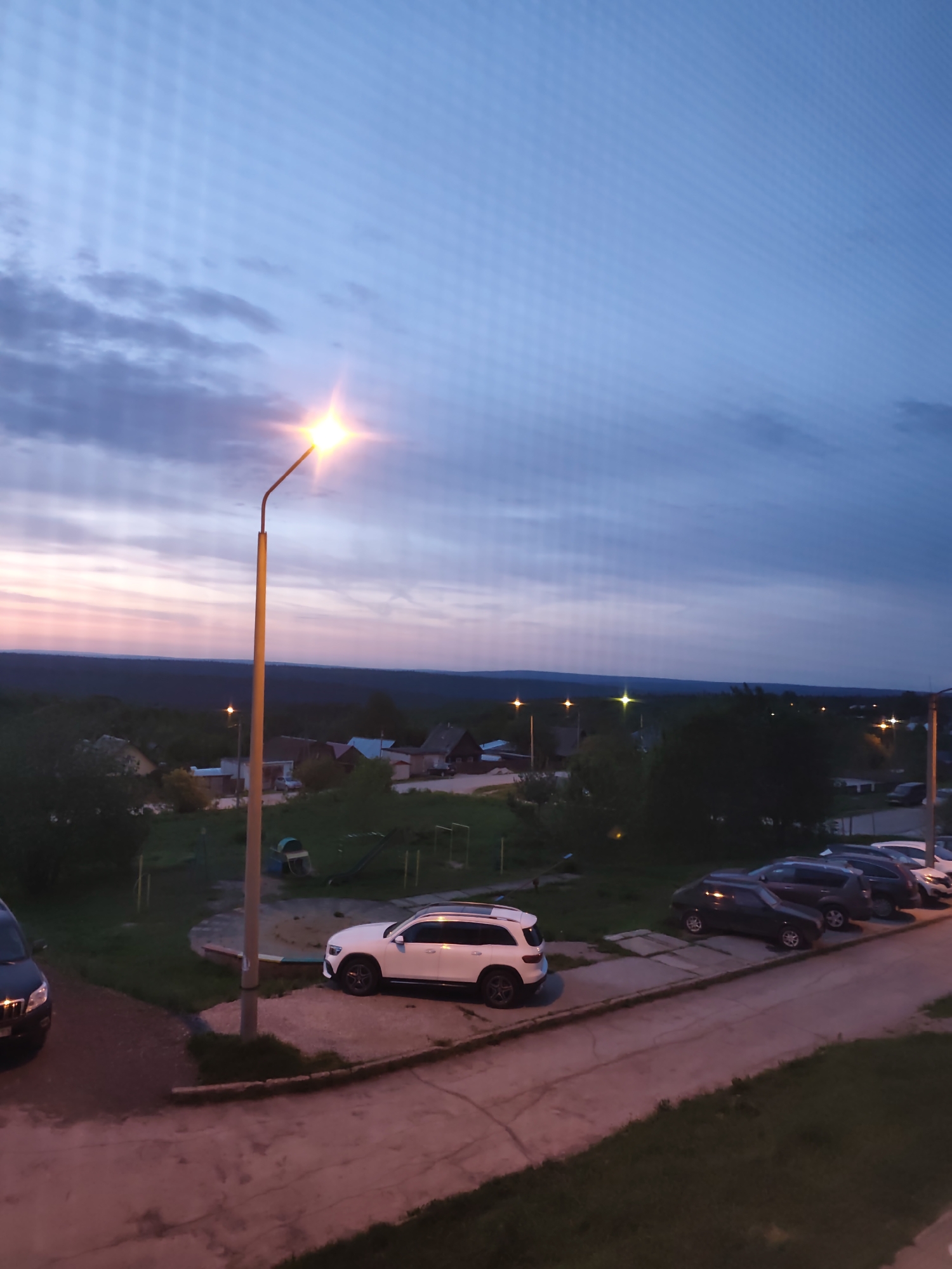 The image depicts a dusk scene in Gremyachinsk, Russia. In the foreground, a street lamp illuminates a parking area along a sidewalk. Several cars are parked, including a white SUV prominently positioned near a playground with a sandpit and slide. Beyond the parking area, the landscape descends to reveal rooftops of houses in the lower ground of the city. In the background, there's a dark, dense forest, possibly marking the edge of the inhabited area. The sky is a gradient of colors, transitioning from orange and pink near the horizon to blue and gray towards the upper part of the image, suggesting either sunrise or sunset. Other street lamps are sparsely distributed in the distance, indicating other residential areas. The area appears residential and quiet.