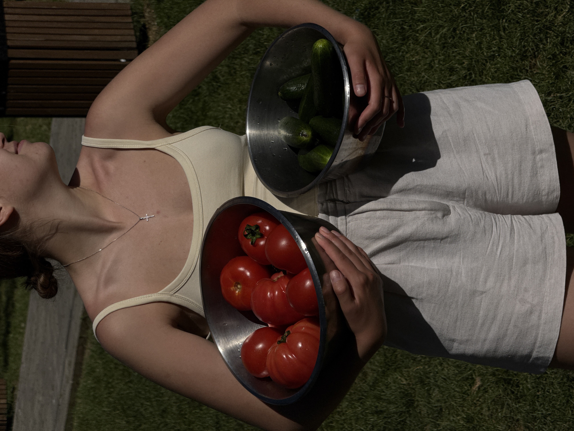 A young woman lies on a grassy surface with interspersed wooden planks. She wears a beige tank top and light-colored shorts. In her hands, she holds two metal bowls: one contains green cucumbers, the other red tomatoes. She is wearing a silver necklace with a small cross pendant. The image suggests a sunny outdoor location, possibly a garden. The city location in the city of Cukarica Urban Municipality, Serbia is not visually determinable from the image.