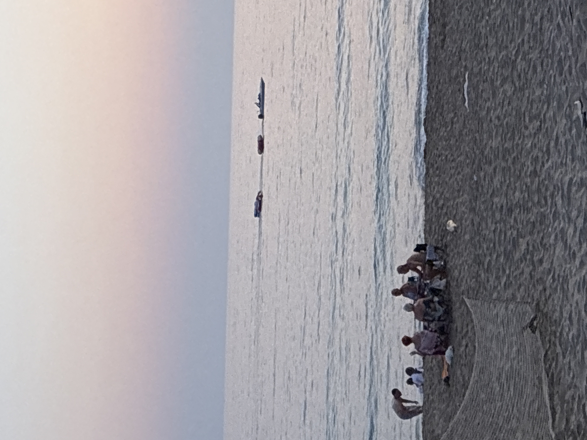 An outdoor, low-angle, full shot captures a beach scene in Alanya, Turkey, during dusk. The sky transitions from a pale pink and orange hue near the horizon to a muted lavender and blue overhead, indicating either sunrise or sunset. The sea is calm, with gentle waves lapping the shore.
On the beach, a group of people are gathered. To the left, a solitary male figure in light-colored shorts bends over near the water's edge. Closer to the group, a family or friends are seated in chairs arranged around a central point. Visible individuals include a person with red hair, another with grey hair, and two males. Some are facing each other, suggesting conversation or shared activity.
In the distance, on the water, several objects are visible. A small boat with a white hull and a dark canopy is moving away from the shore. Further out, two inflatable tubes, possibly being towed, are also present on the sea. The beach itself is composed of dark sand, with a fishing net laid out in the foreground, partially obscuring the sand. Scattered items on the beach include what appear to be white plastic bags or debris. The overall atmosphere is serene and tranquil, characteristic of a quiet evening by the sea in Alanya.