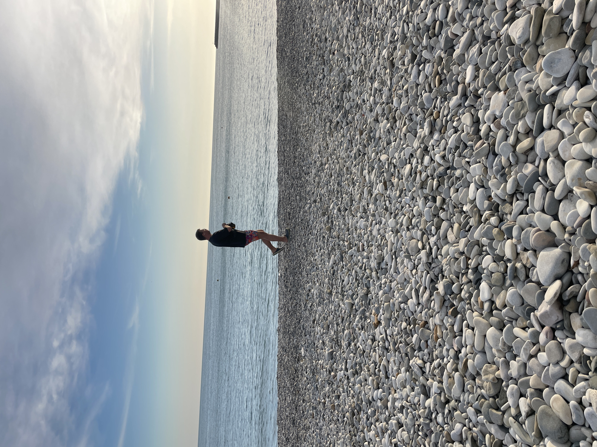 The image depicts a person standing on the edge of a pebble beach along the coast. The person is wearing a dark shirt and patterned shorts. The beach is covered in smooth, gray pebbles. The water is calm and stretches to the horizon. The sky is blue with some clouds near the top of the image. The location appears to be Olginka, Russia.