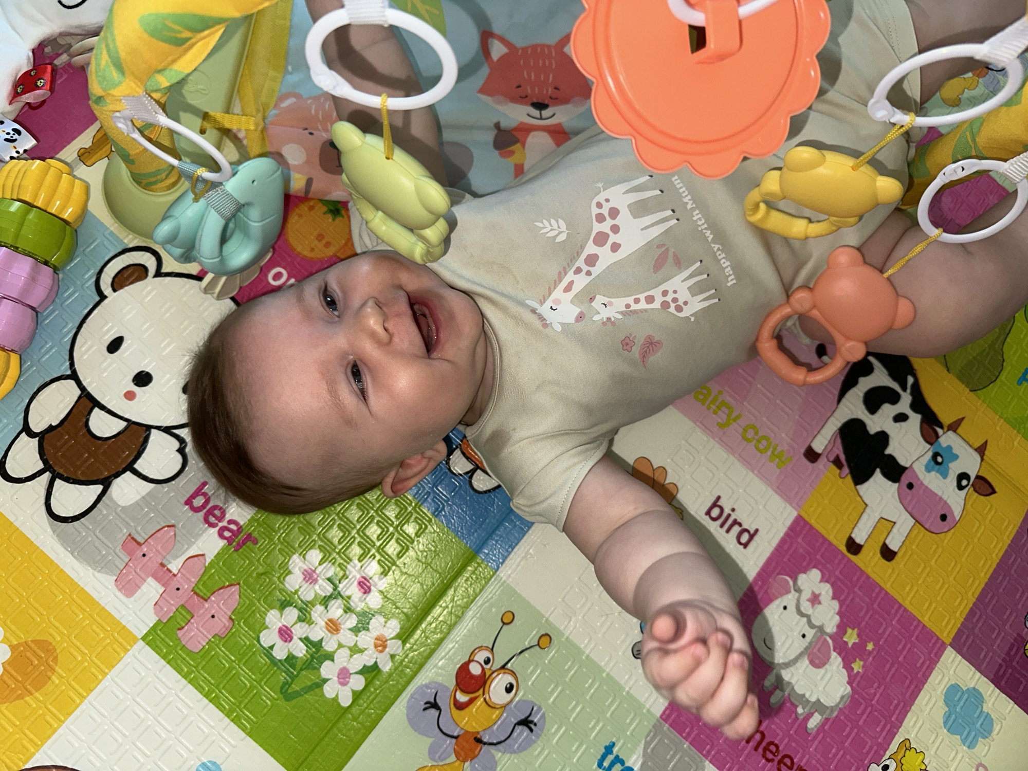 A baby lies on their back on a colorful play mat in what appears to be a domestic setting. The baby is wearing a light-colored onesie with a giraffe pattern and the words "Happy with Mum" printed on it. Several toys hang above the baby, including a green elephant, a yellow bear, and an orange flower shape. The play mat is decorated with animal and plant designs, including a bear, flowers, a bee, and a cow. The baby is smiling and appears to be engaging with the toys. The location is Chaikovsky, Russia.
