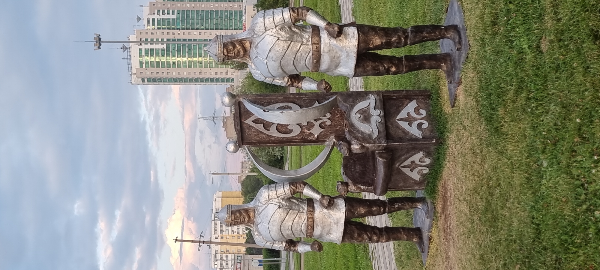 The image depicts an outdoor monument in Aktobe, Kazakhstan, featuring two warrior statues standing on either side of a decorative throne. The statues are made of metallic material and are clad in armor with helmets. The throne is adorned with intricate patterns and a crescent moon symbol. In the background, modern high-rise buildings and power lines are visible, indicating an urban setting. The ground is covered with green grass and paved pathways. The sky is partially cloudy with soft lighting, suggesting it is either early morning or late afternoon.