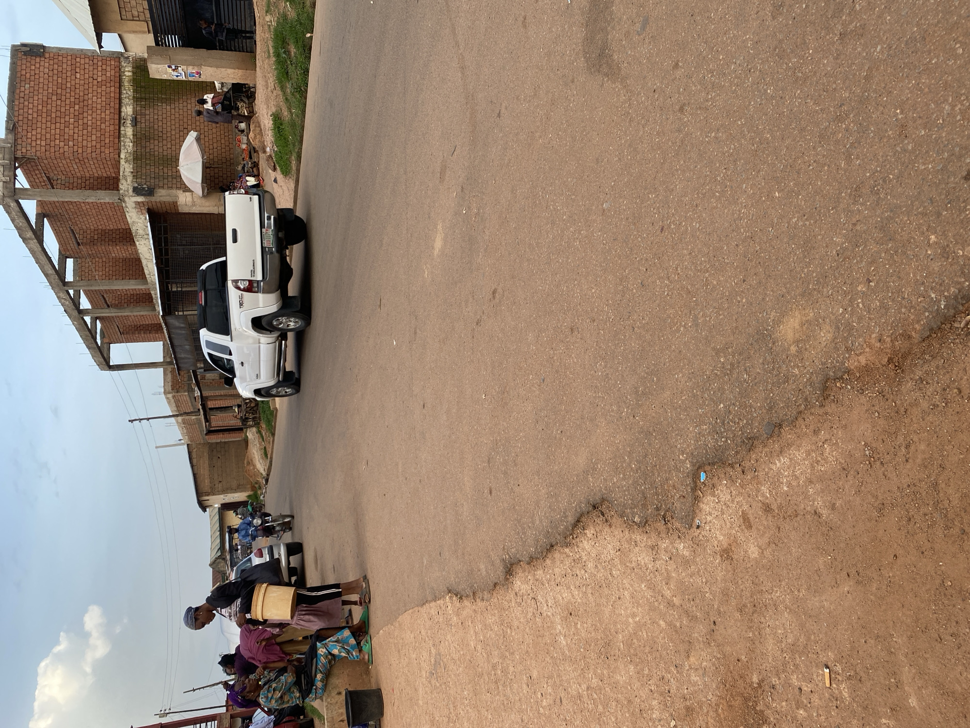 In Bukuru, Nigeria, a white Toyota Tacoma pickup truck is parked on the right side of a paved road. The truck is positioned on the driving lane, facing forward. In the background, a partially constructed brick building stands with exposed rebar. To the left of the road, several individuals are gathered on a dirt shoulder. One woman is seated on a raised concrete curb, wearing a blue and green patterned dress. Another individual, wearing a black top and leggings, stands holding a large yellow bucket. Further down the road, a man rides a motorcycle, and other figures are visible near market stalls under a striped umbrella. The sky above is partly cloudy.