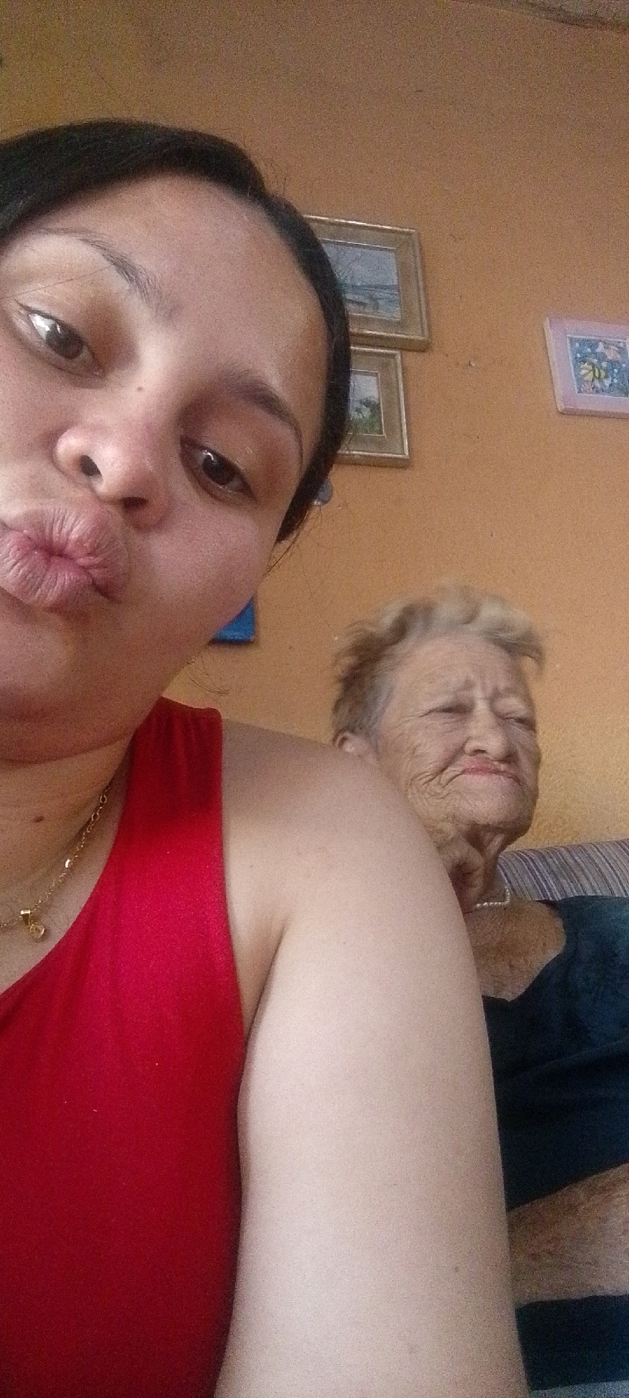 A selfie is taken by a woman with dark hair and brown eyes, puckering her lips. She is wearing a red top and a gold necklace. In the background, an elderly woman with light-colored hair is visible, with her eyes closed and a slight smile. She is wearing a dark top and a pearl necklace. The setting appears to be indoors, with an orange-tinted wall and framed pictures hanging on it. The image quality is grainy. This photograph was likely taken in Juan Griego, Venezuela, a coastal town known for its historical fortifications and vibrant atmosphere.