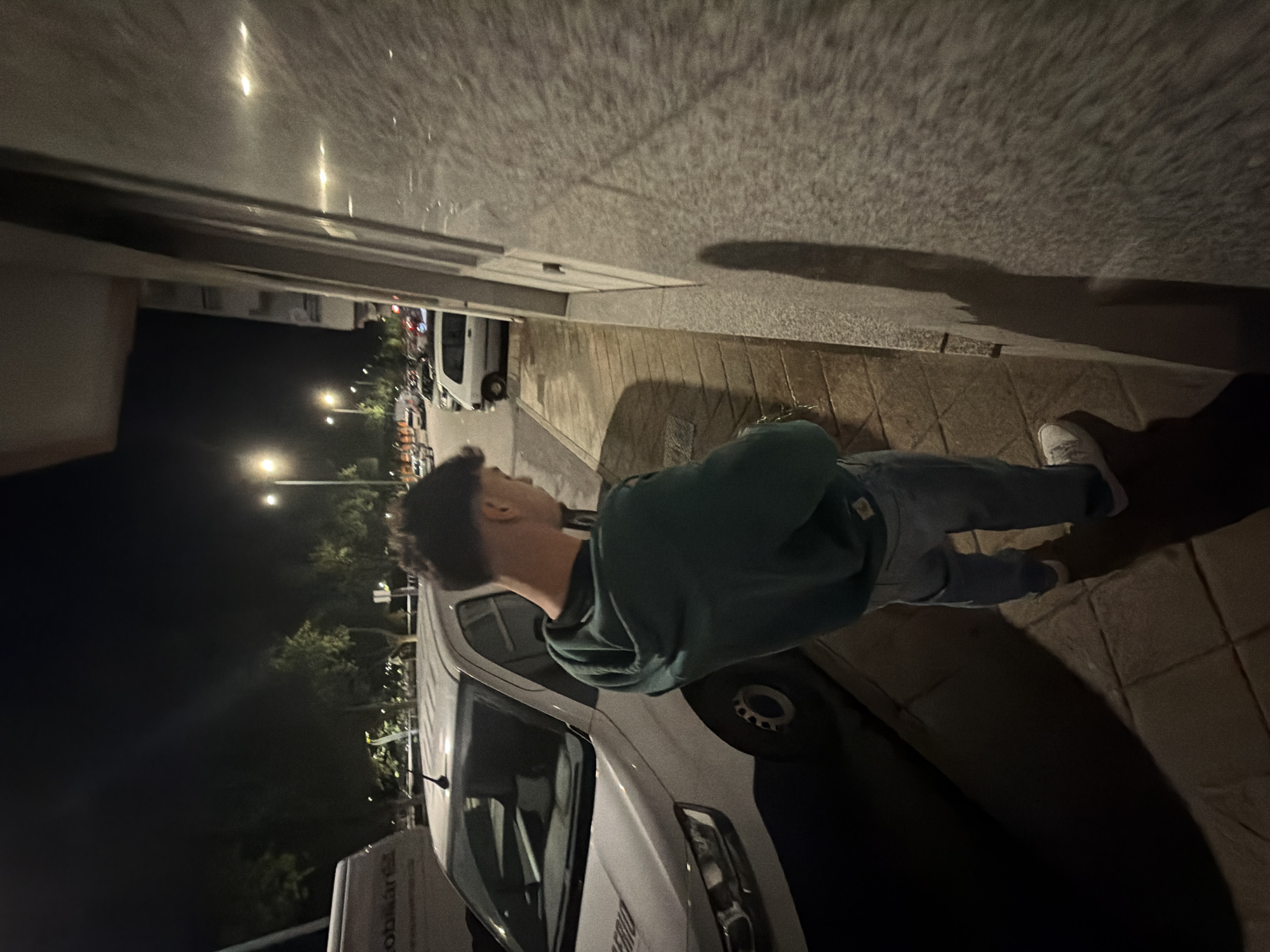 The image shows a man standing on a sidewalk at night in Bragança, Portugal. He is wearing a green hoodie, jeans, and white sneakers. He is standing next to a white van parked on the street. Streetlights illuminate the scene. There are other parked cars and buildings visible in the background. The wall of a building is on the right side of the frame, with the man's shadow cast on it.