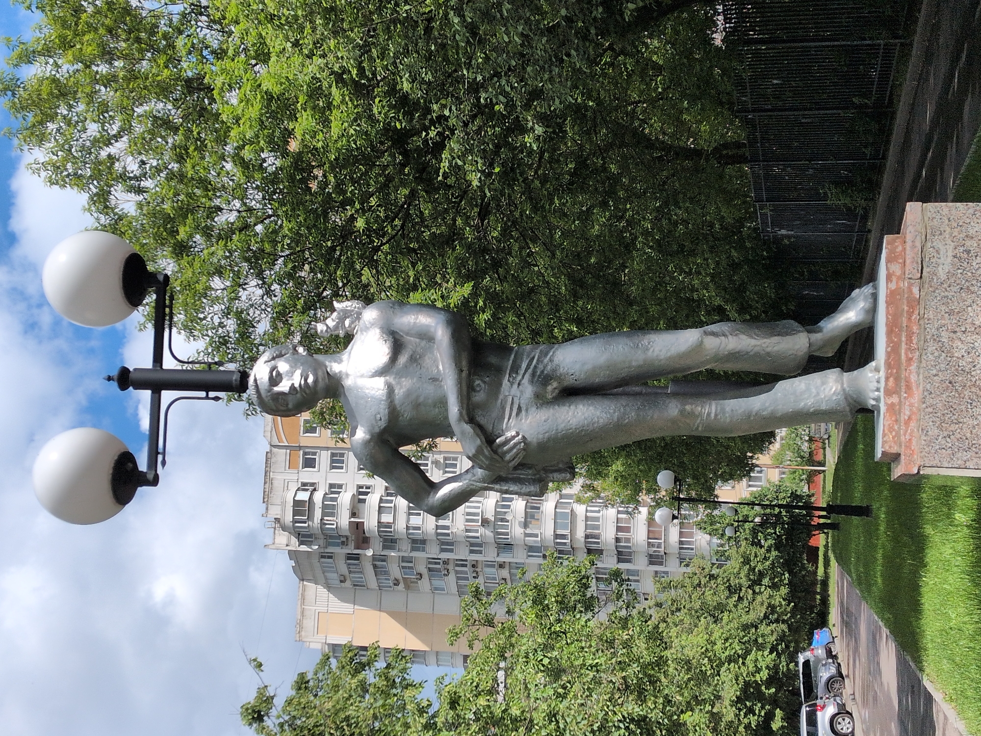 The image depicts a silver-colored statue of a reclining male figure in a park-like setting. The statue is positioned atop a granite base, with a mature green tree providing a backdrop. To the left of the statue is a black lamppost featuring two spherical white lamps. In the background, a large, multi-story apartment building with numerous windows is visible, suggesting an urban residential area. A stretch of green lawn extends towards the right edge of the frame. In the distance, another lamppost and parked cars can be seen. The location is likely Moscow, Russia.