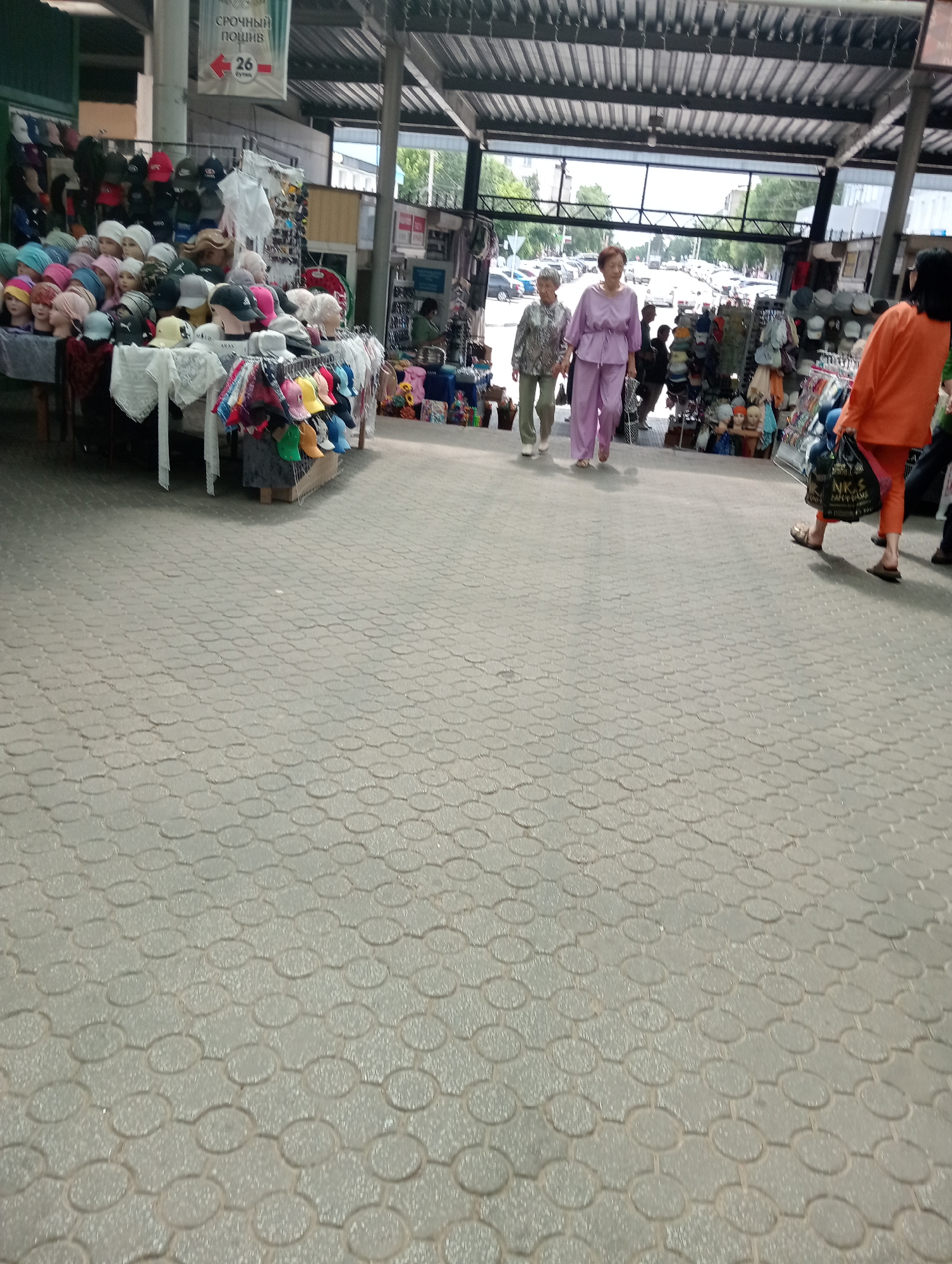 The image depicts an outdoor marketplace in Kostanay, Kazakhstan. In the foreground, a textured pavement is visible. Several individuals are present. Two older women are walking towards the left side of the frame. One is wearing a patterned blouse and light-colored pants, while the other is in a lavender pantsuit. To the right, a person in an orange shirt and pants is walking away from the camera, carrying a dark bag. In the background, stalls display various items, including hats, clothing, and other merchandise. The marketplace appears to be covered by a metal roof structure. The scene suggests a typical day at a local market with shoppers and vendors.