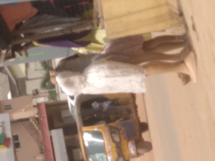 This image captures a street scene in Agege, Nigeria. In the foreground, positioned to the right, two individuals are partially visible, exhibiting a soft focus. One individual wears a light-colored, possibly white, patterned dress reaching mid-thigh, complemented by light-toned flat shoes. Adjacent and partially behind, a second individual is discernible in a light purple or pinkish dress. They are situated next to a structure constructed with wooden supports, from which various colored fabrics—including purple, yellow, and orange—are suspended, indicative of a market stall. The ground in this area is unpaved and appears as a light brown surface.
In the midground, to the left, a yellow and blue Keke Napep (auto-rickshaw) is present, with two indistinct figures visible within its passenger compartment. A white vehicle is partially visible behind the Keke Napep. The background reveals multiple buildings: one features a prominent multi-colored geometric sign (red, green, blue, yellow) on its upper facade, and another building exhibits a reddish hue with an external staircase. The environment suggests an active urban or commercial setting.