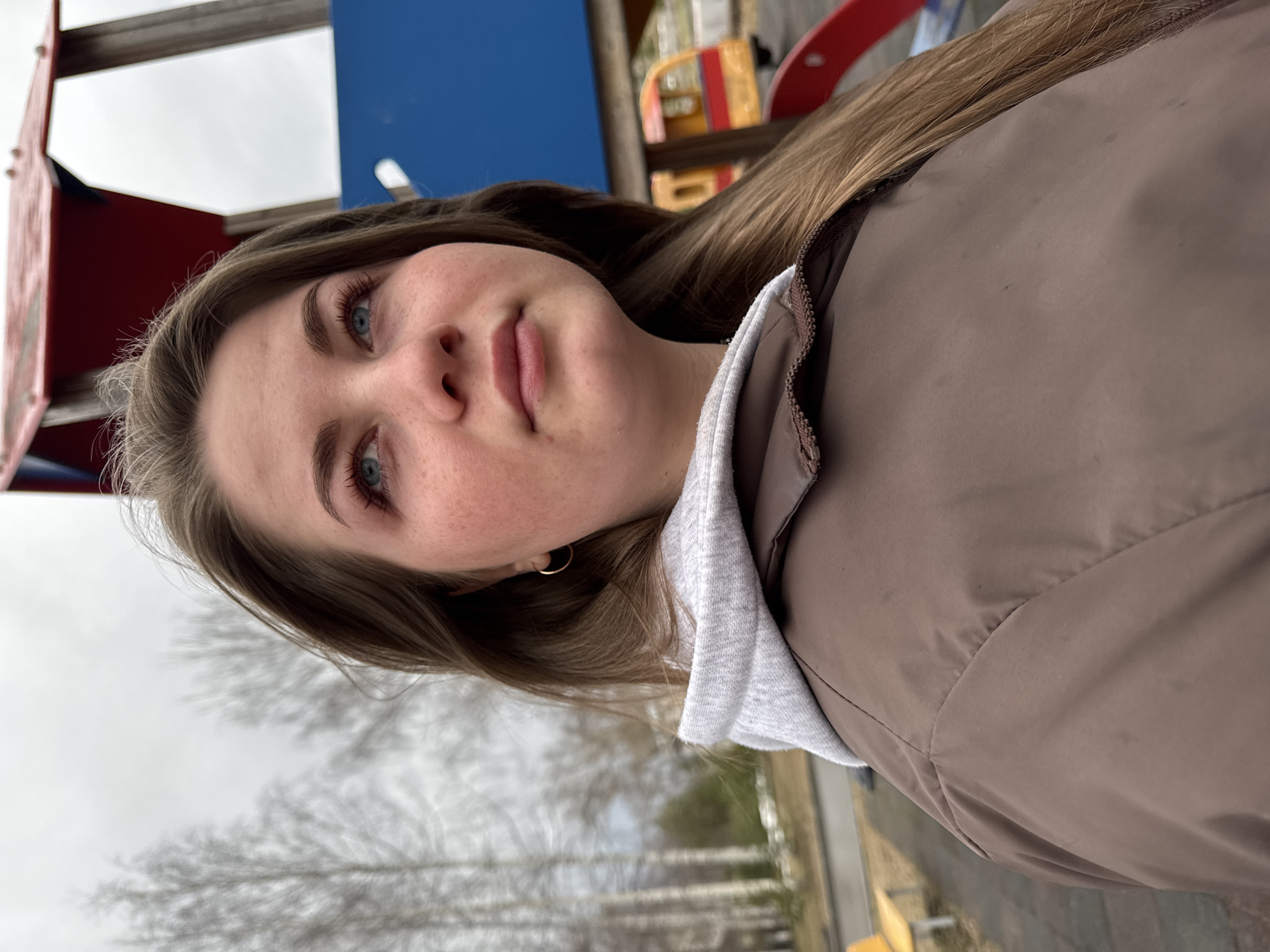 A close-up, medium-shot photograph shows a young woman with light brown hair and blue eyes looking at the camera. Her fair skin is slightly flushed. She wears a brown jacket with a gray hoodie underneath.
The background reveals a playground structure with red, blue, and yellow elements. Bare trees are visible, suggesting a fall or winter setting. The location is the city of Chaikovsky, Russia.