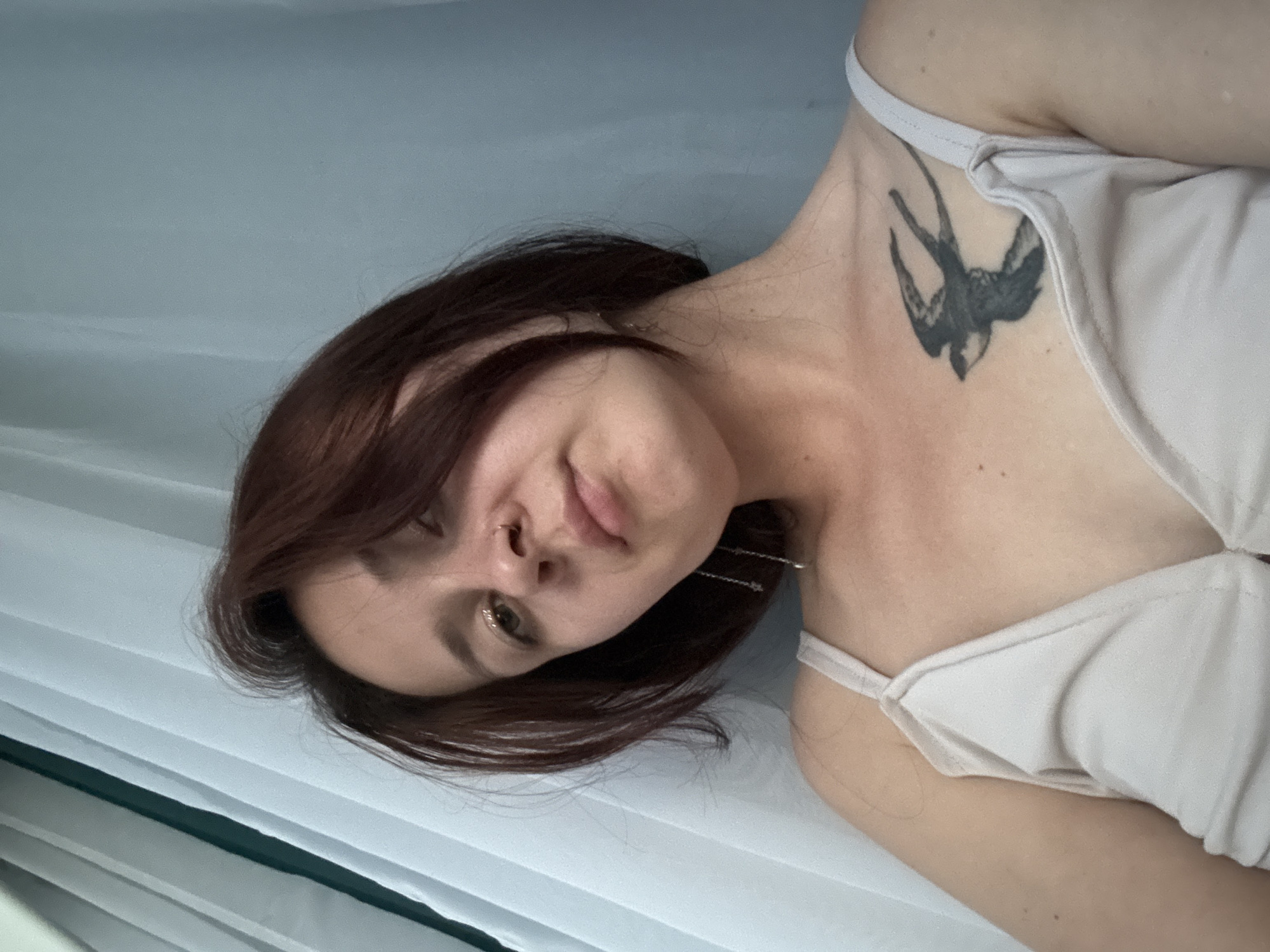 A young woman with short, dark brown hair and a piercing on her nose looks directly at the camera. She is wearing a light-colored tank top, and a tattoo of a bird is visible on her chest. She has glittery eyeshadow on her eyelids. She is leaning against a light-colored, sheer curtain. The scene is likely indoors, possibly in an apartment, in Naberezhnye Chelny, Russia.