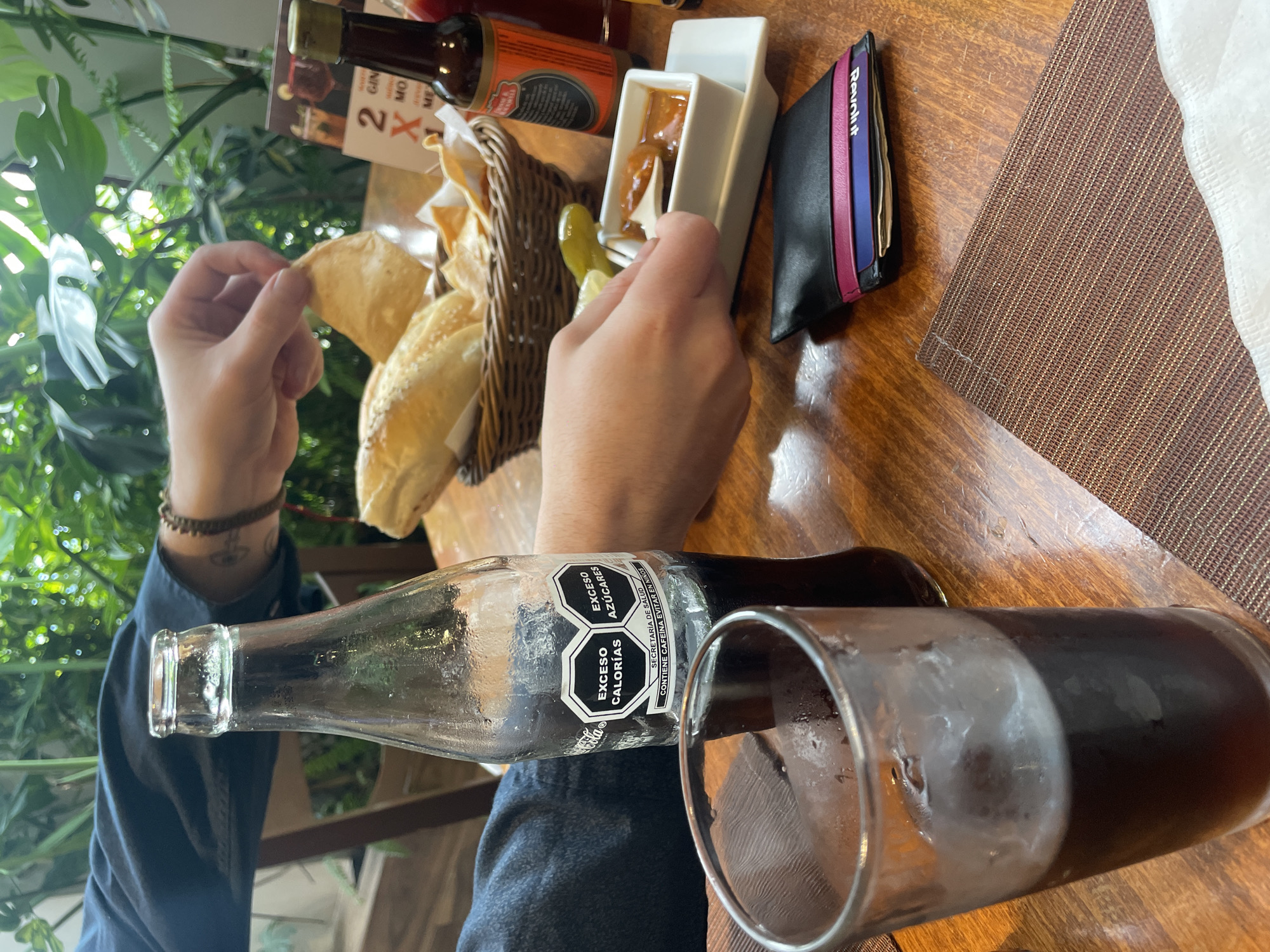The image depicts a close-up view of a table at an outdoor restaurant, likely in Cuautitlán Izcalli, Mexico, with lush green foliage in the background. A glass of dark beverage with ice is prominently featured in the foreground, next to a glass bottle of Coca-Cola. The bottle's label displays nutritional warnings in Spanish, indicating "EXCESO CALORIAS" (Excess Calories) and "EXCESO AZUCARES" (Excess Sugars).
In the middle ground, a hand is shown reaching into a basket of tortilla chips and bread. Beside the basket is a white dish containing a dipping sauce and a slice of pickled jalapeño pepper. A small wooden utensil is being used to scoop the sauce. To the right of the dipping sauce, a small bottle of hot sauce with a red label is visible. A black wallet with colorful card slots is placed on the table near the dipping sauce.
The table is made of wood, and a textured brown placemat is partially visible in the lower right corner. The overall scene suggests a casual dining experience, possibly involving appetizers or snacks. The presence of specific food items and the Coca-Cola bottle with Mexican labeling strongly indicate a Mexican setting. The natural greenery in the background implies an open-air or well-lit interior space, perhaps a garden restaurant.