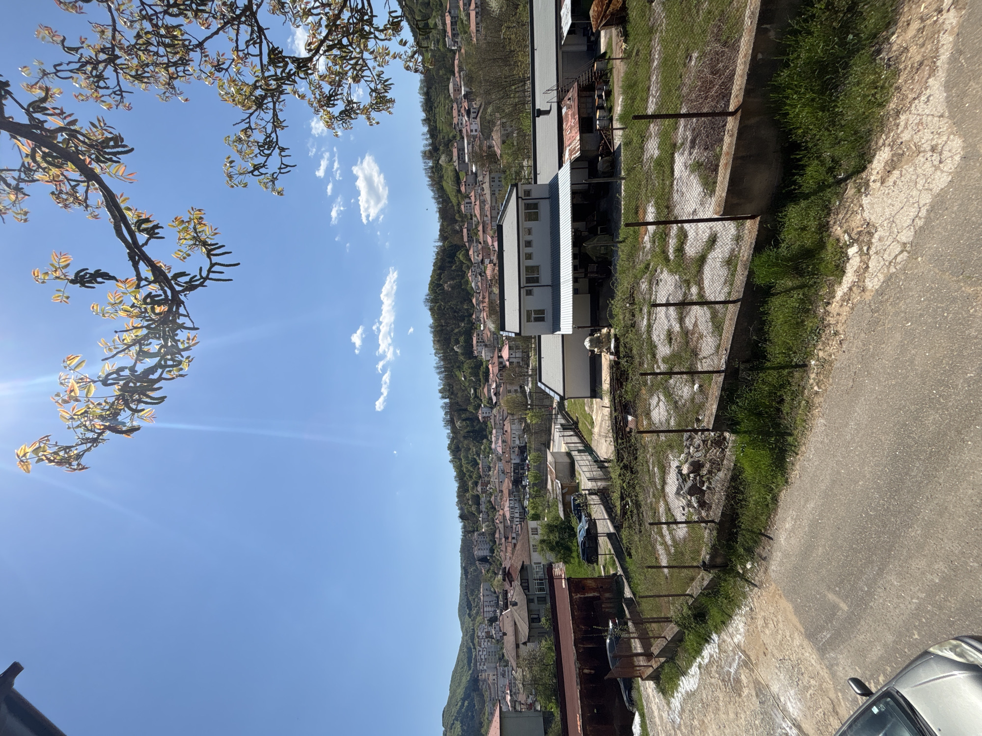 The image depicts a neighborhood in Zlatograd, Bulgaria, taken from an elevated position. The scene features a blue sky with scattered clouds, a tree branch with budding leaves in the upper left corner, and a hillside dotted with houses. A fence runs along the right side of the frame, bordering a concrete pathway and road. Adjacent to the fence are structures, including a multi-story building and other constructions. Part of a silver car is visible in the bottom right corner. The overall environment suggests a residential area nestled in a hilly landscape.