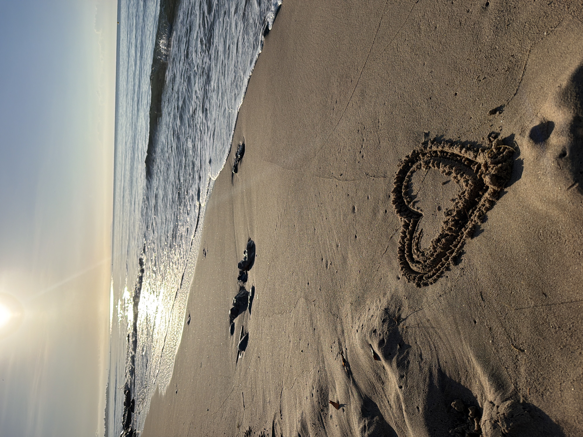 The image depicts a tranquil beach scene at sunrise in Mont-roig del Camp, Spain. A heart shape has been drawn in the wet sand near the waterline. Gentle waves are lapping at the shore, and the sun is low in the sky, casting a warm, golden light and creating reflections on the water. The sand is a light brown color, with some ripples and footprints visible. In the distance, the coastline stretches out, and the sky is a pale blue. There are no people visible in the frame.