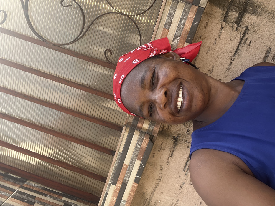 An adult female is depicted in a medium-shot, facing directly towards the camera with a broad smile, her teeth visible. She has a medium complexion and wears a red bandana with white patterns covering her head, a royal blue sleeveless top, and a dark blue teardrop-shaped earring with a white accent on her right ear. The background, from bottom to top, features a tan textured wall, followed by a horizontal band of multi-colored rectangular tiles in brown, beige, and black hues. Above the tiles, a translucent corrugated or ribbed structure, possibly a window or awning, is visible, supported by diagonal dark-colored bars. Part of a decorative dark metal scrollwork, consistent with security grating, is present on the upper right. The scene is brightly lit, indicative of an outdoor environment in Iranla, Nigeria.