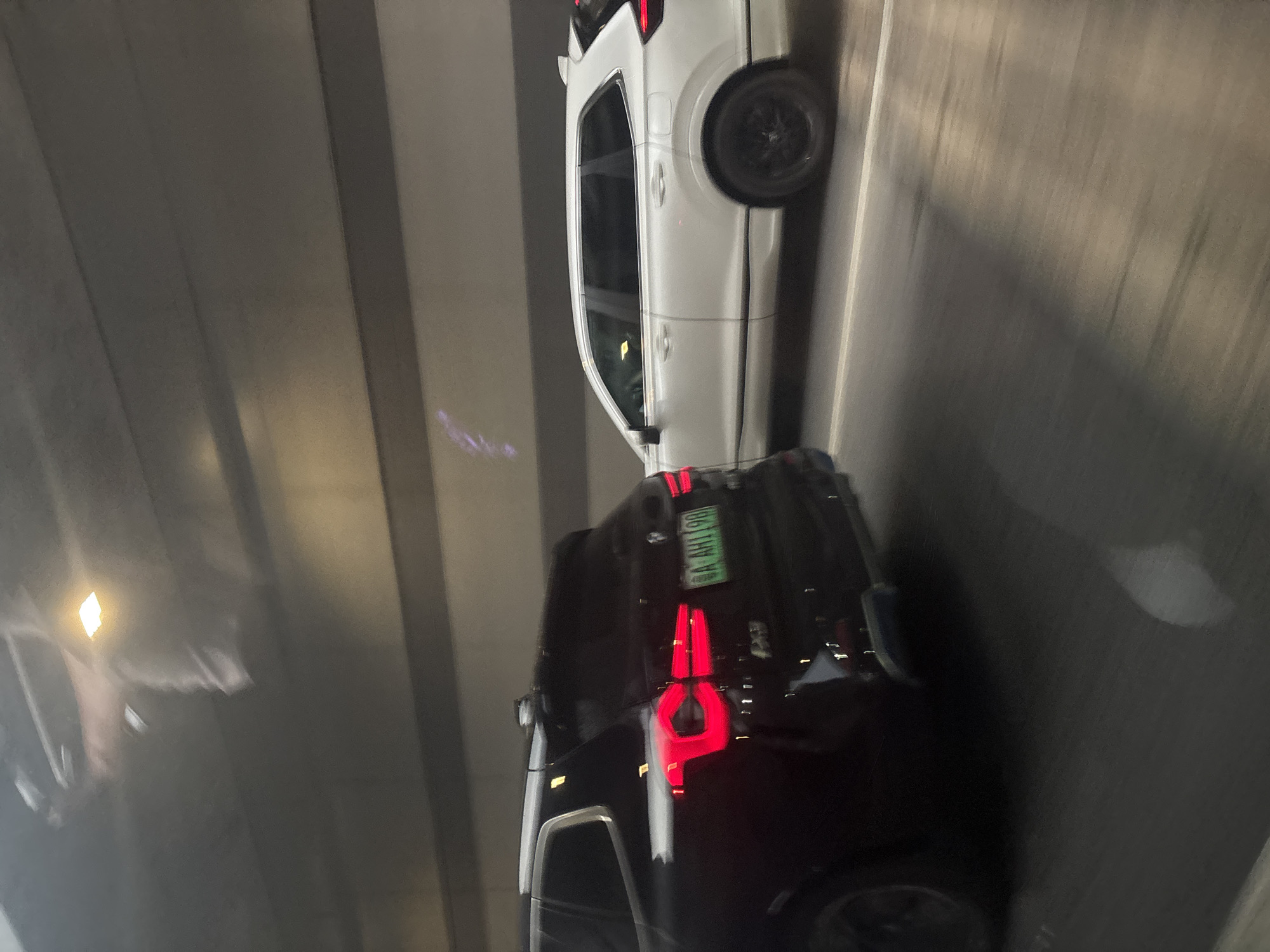 The image depicts two SUVs in what appears to be a tunnel or underpass in Tianhe District, China. The foreground features a black SUV, facing away from the camera, with its taillights illuminated. A green license plate with characters "A AHI 19" is visible on the back. To the right of the black SUV is a white SUV, also facing away, its side profile clearly visible. Both vehicles are in motion, as indicated by the motion blur on the road and surrounding environment. The tunnel walls are a muted gray with horizontal lines, and overhead lighting is present. There are no people clearly visible within the vehicles or in the immediate vicinity.
