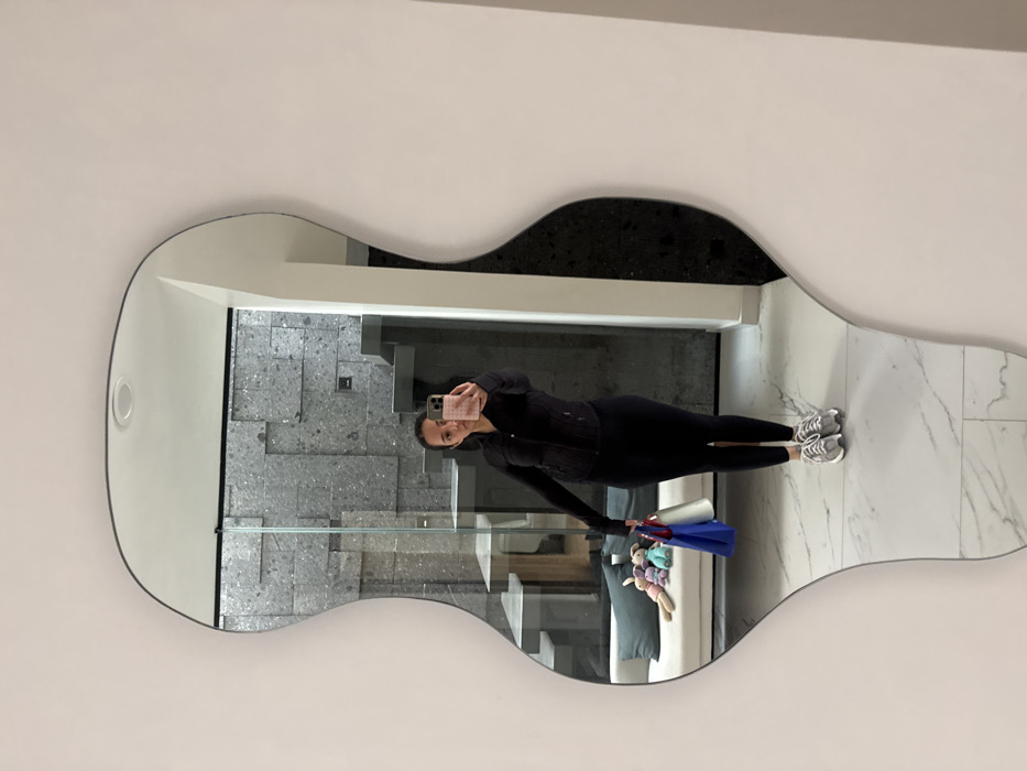 A woman wearing a black athletic outfit takes a mirror selfie. The mirror has an unusual, guitar-like shape and is hanging on a white wall. The reflection shows a hallway with gray tiled stairs, a glass railing, and a white marble floor. The woman holds shopping bags and a stuffed animal.
