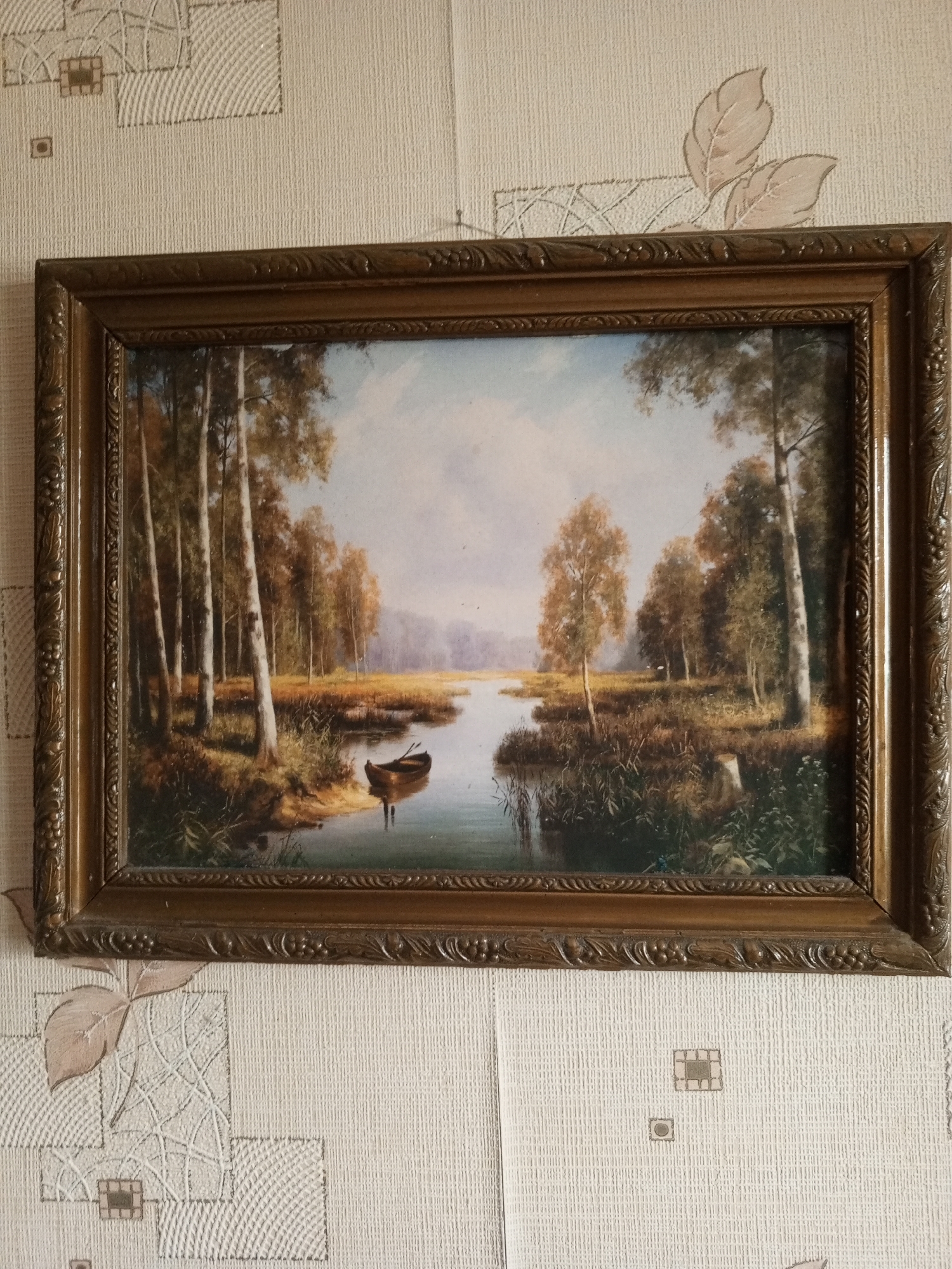 A framed oil painting depicts a serene natural scene. The artwork showcases a tranquil body of water with a small, traditional wooden rowboat adrift near the shore. Tall, slender trees with autumnal foliage line the bank, their reflections subtly mirroring in the calm water. The landscape extends to a distant, hazy horizon, suggesting a vast, open natural environment. The overall mood is peaceful and contemplative. The painting is framed in an ornate, dark-colored frame, possibly made of wood, with intricate carvings. The artwork is hanging on a textured wallpapered wall. The wallpaper has a repeating pattern of faint floral motifs and geometric shapes in a light, neutral color. The scene likely represents a quiet moment in a rural or natural setting, perhaps a lake or slow-moving river in a wooded area. The time of day appears to be late afternoon or early morning, given the soft, diffused light and the muted colors of the sky and landscape. There is no visible text in the image. The provided location context is Nizhny Novgorod, Russia.